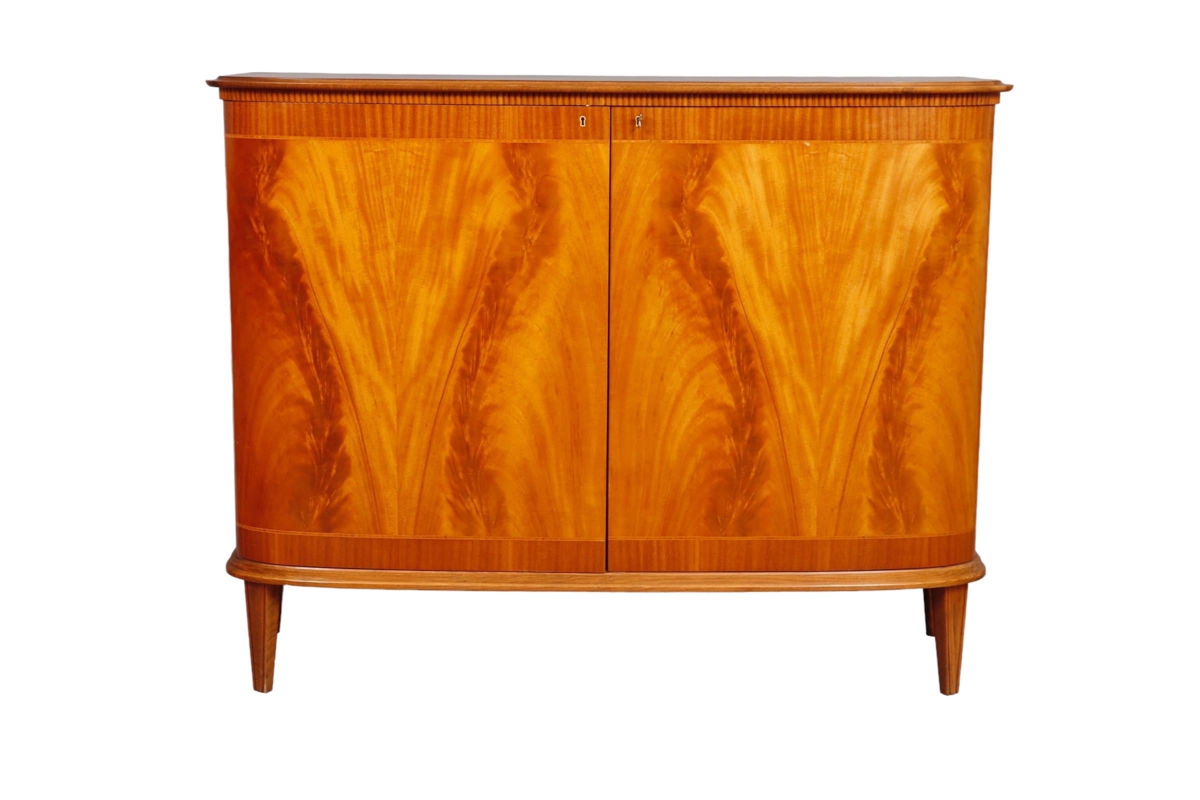 1940’s sideboard by Swedish company Skänk. Stunning flame mahogany veneers decorate inlaid bentwood cabinet doors. Inside are three shelves and two small drawers.