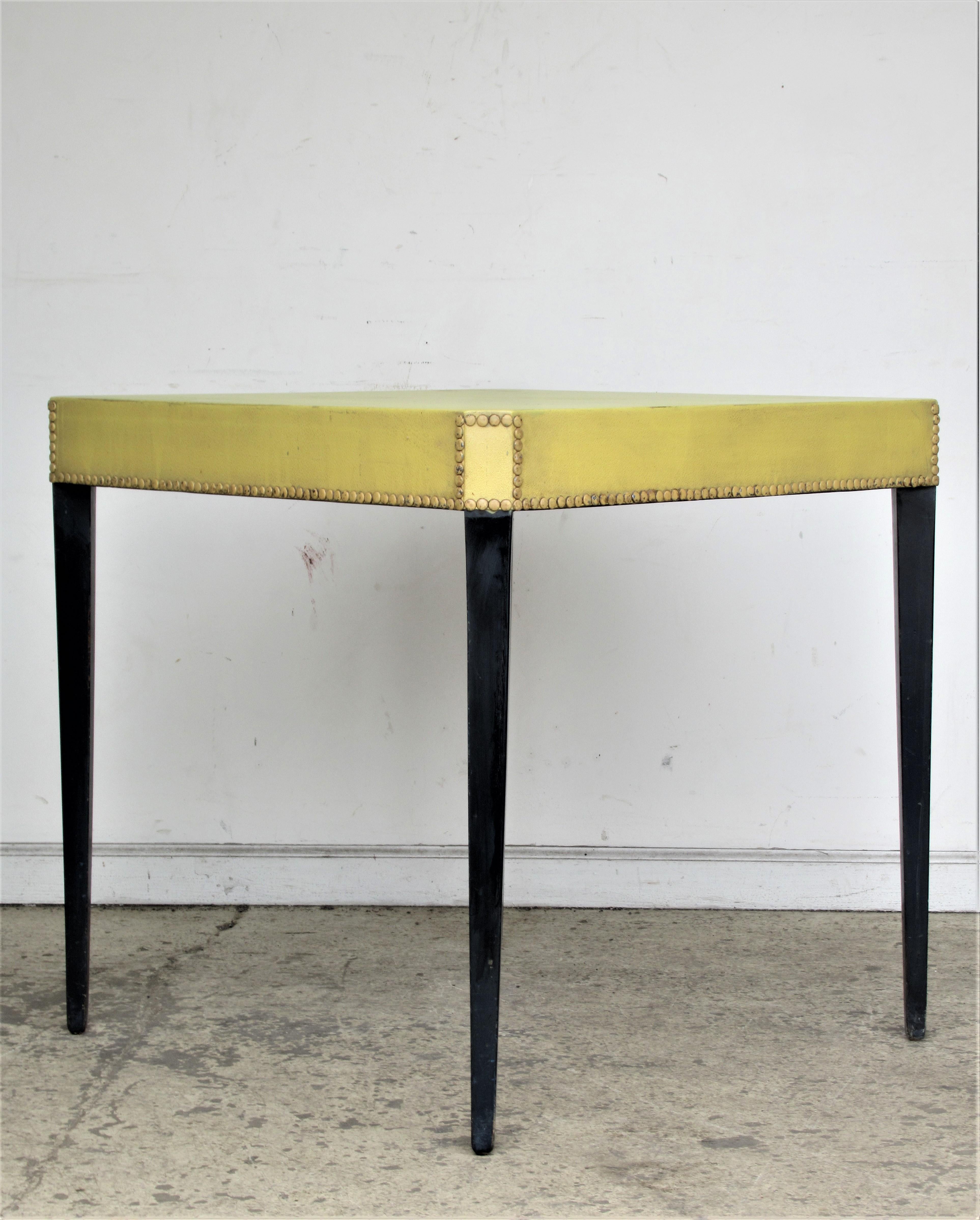 Sleek 1940s occasional table in the style of Tommi Parzinger with original ebony painted tall tapered wood legs and the original yellow paint stained textured vinyl top fully surrounded by brass tacking on all sides. Old paper label on underside -