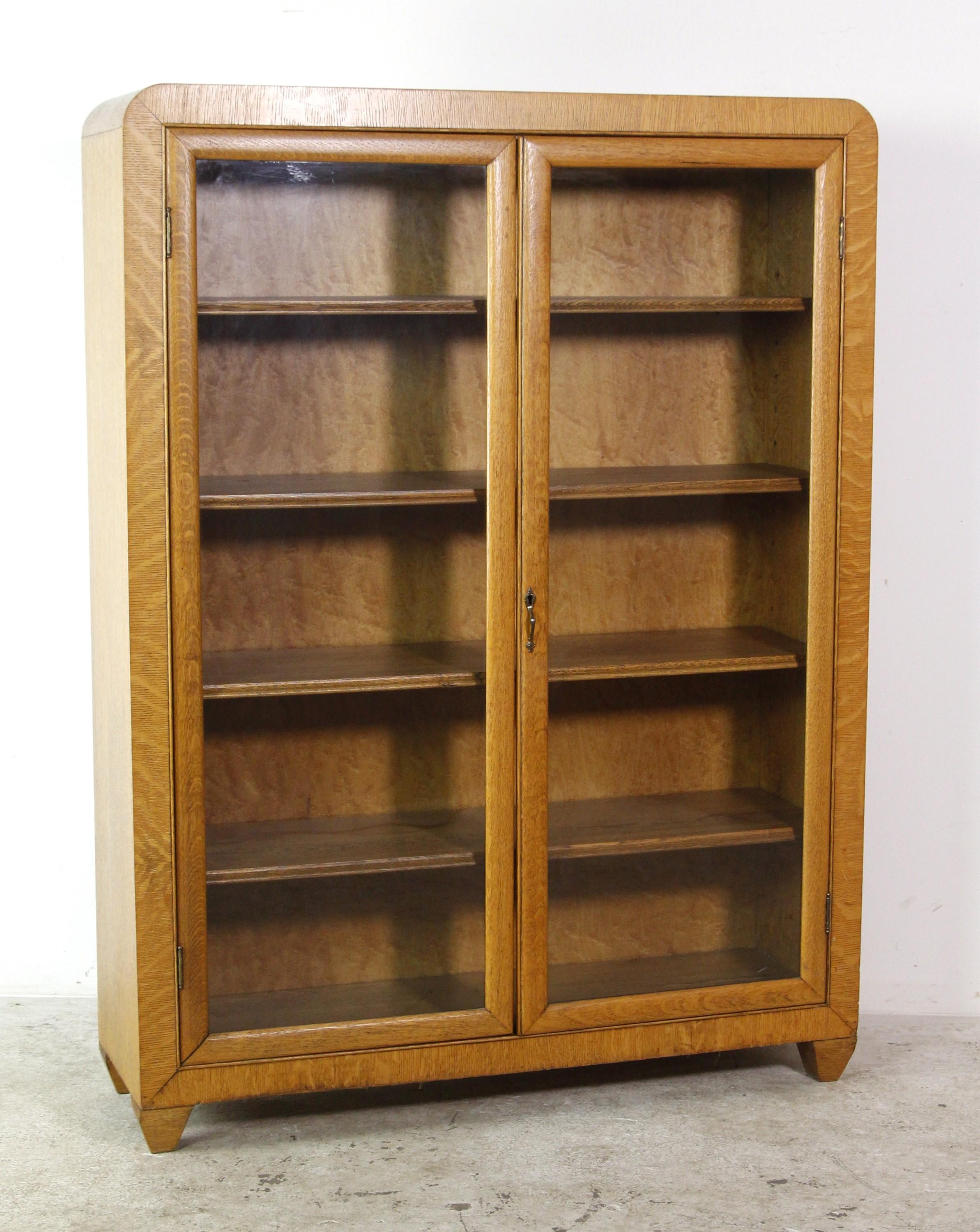 1940s Mid-Century Modern tiger oak bookcase cabinet with two front glass doors and four shelves.  It includes the original brass handle and key. There is some minor damage to the back. Please see the photos. This can be seen at our 400 Gilligan St