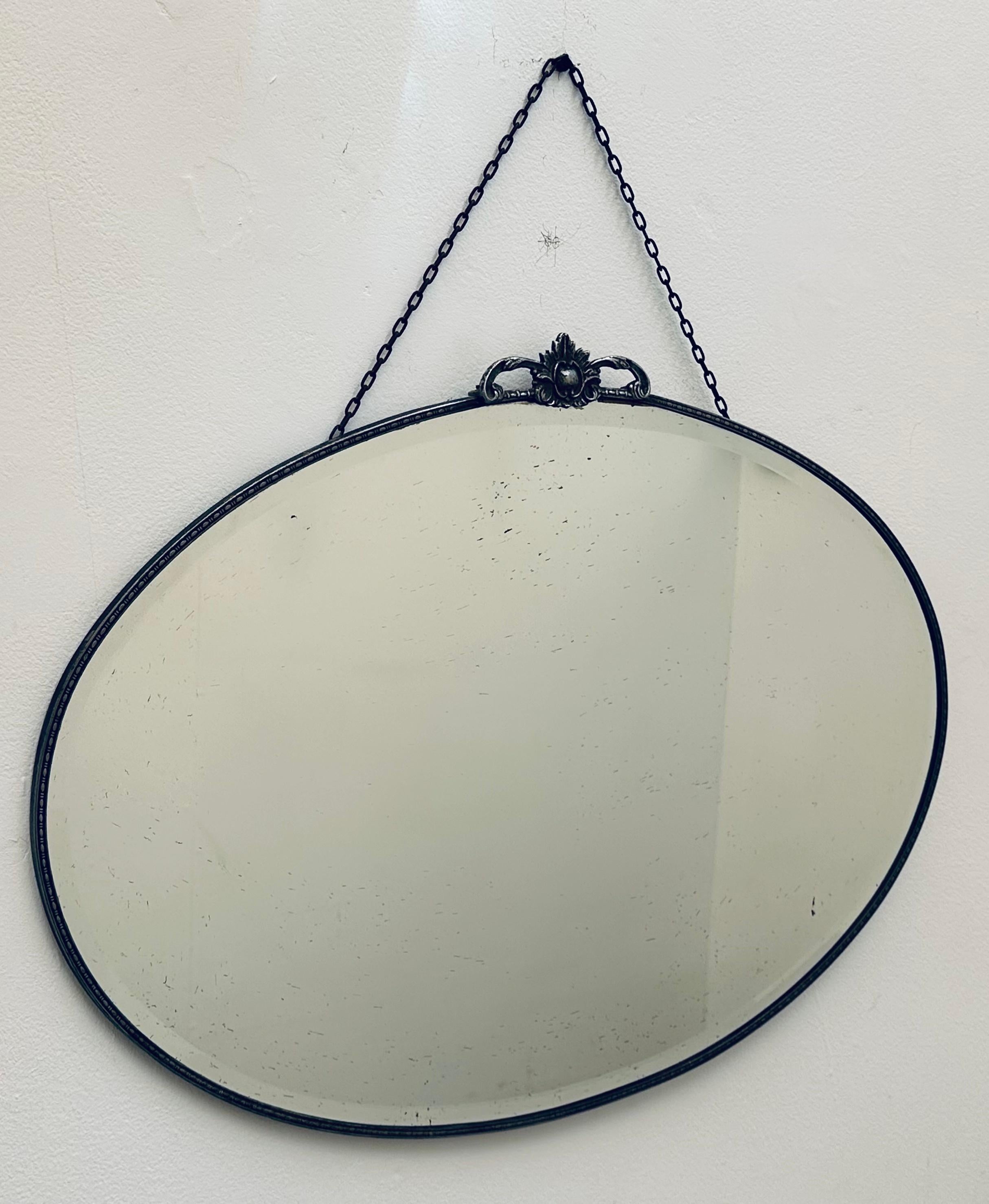 A large 1940s bevelled-edge oval glass and chrome mirror.  A flower shaped chrome crest sits at the top with a thin chrome frame running around its circumference.  A rusty linked hanging chain is fixed to the back of the top of the mirror allowing