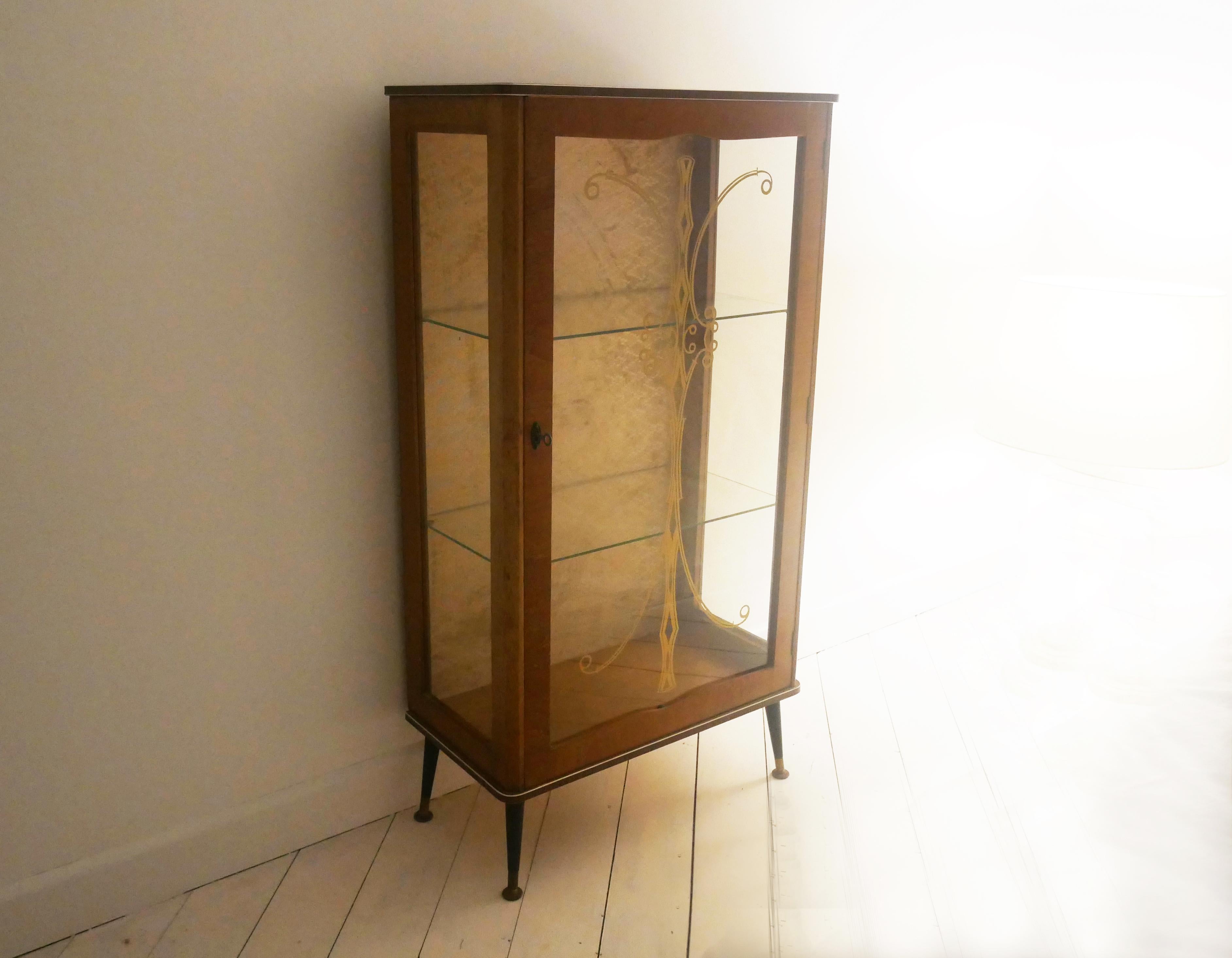 Elegant 1950’s vintage glass display cabinet, with gold decoration on door, and two glass shelves within. The interior back is lined with the original silver patterned material. Sits on black painted teak las with brass tips. Key supplied.

Size