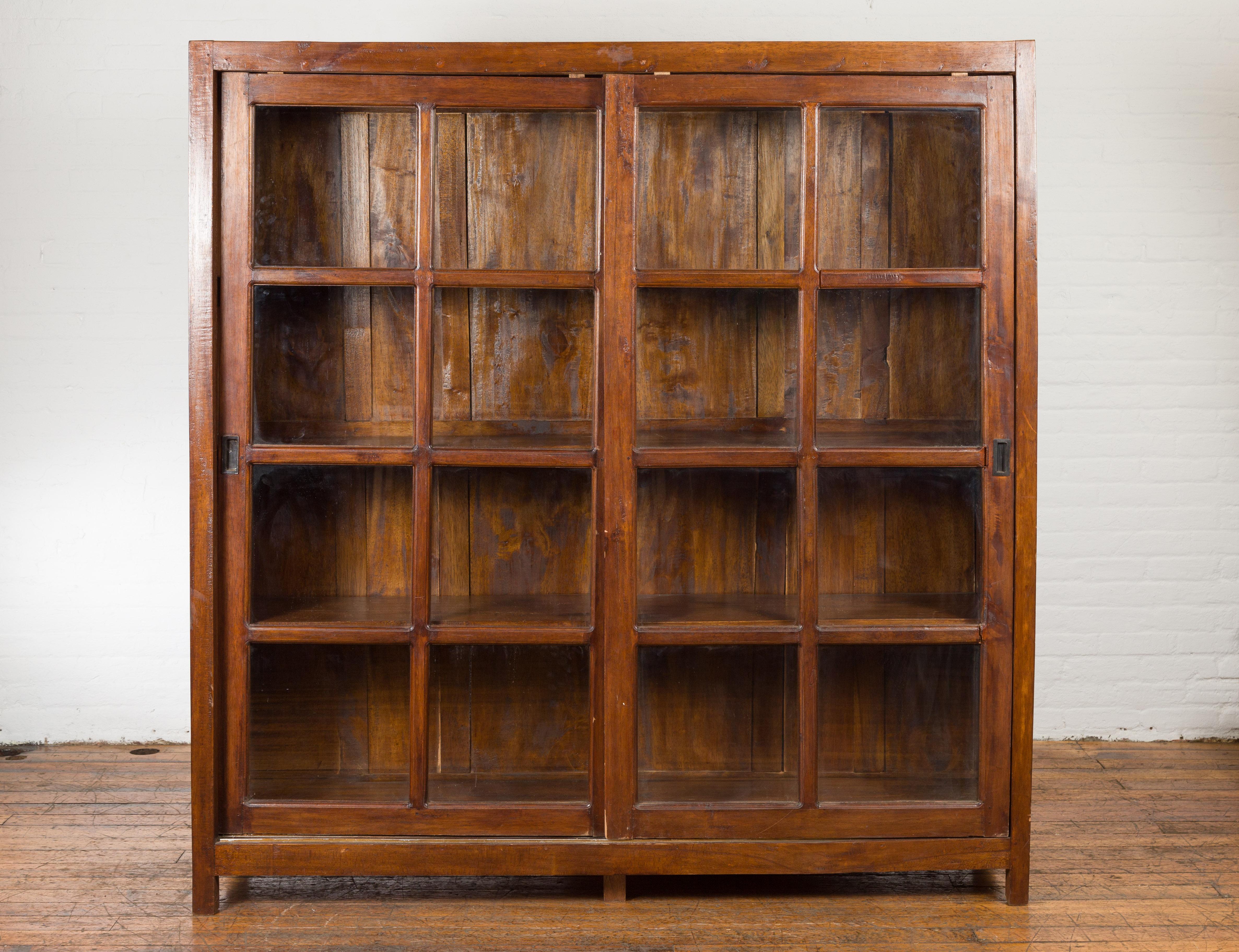 A Javanese wooden vitrine bookcase from the Mid-20th Century, with sliding paneled glass doors and brown patina. Created on the Island of Java during the second quarter of the 20th century, this wooden bookcase features a linear silhouette perfectly