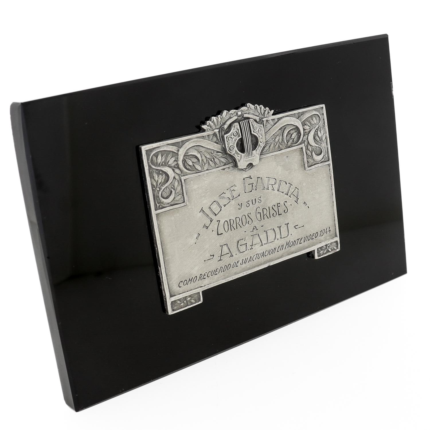 Una bella targa commemorativa antica.

Con una targa in argento (o bronzo argentato) posta su una base di vetro nero.

La targa è dedicata con l'iscrizione: 