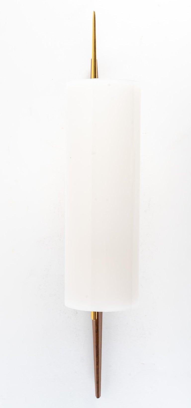 Composé d'un support mural rectangulaire en laiton doré sur lequel repose un long bras conique en bois de rose.
Au centre du bras est positionnée une rotule en laiton doré sur laquelle est posée une longue tige en laiton doré ornée d'un grand