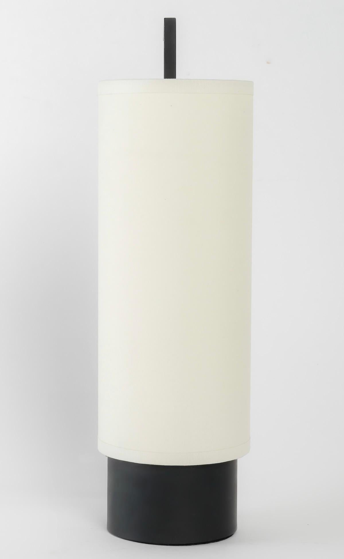 Composé d'un socle cylindrique noir sur lequel repose un abat-jour en coton blanc cassé de forme cylindrique épousant le socle et maintenu par une tige en fer forgé noir de section carrée qui longe l'abat-jour et se prolonge vers le haut à angle