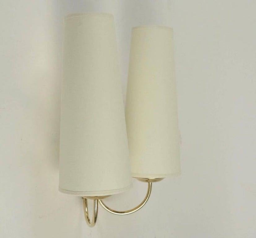 Pair of 1950s Arlus brass sconces.

Composed of a wall support in gilded brass receiving in the lower part two rising rods placed on either side of the gilded brass wall lamp. 
They are dressed with conical lampshades in off-white cotton and