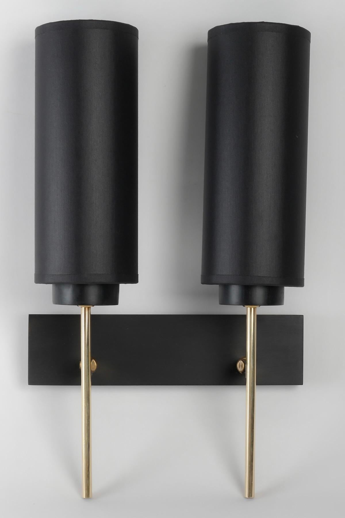 Composed of a rectangular wall bracket in blackened wood on which rest two gilded brass light arms positioned vertically on the bottom of the sconce.
On the upper part, the light arms are clad with cylindrical black cotton shades, highlighted by