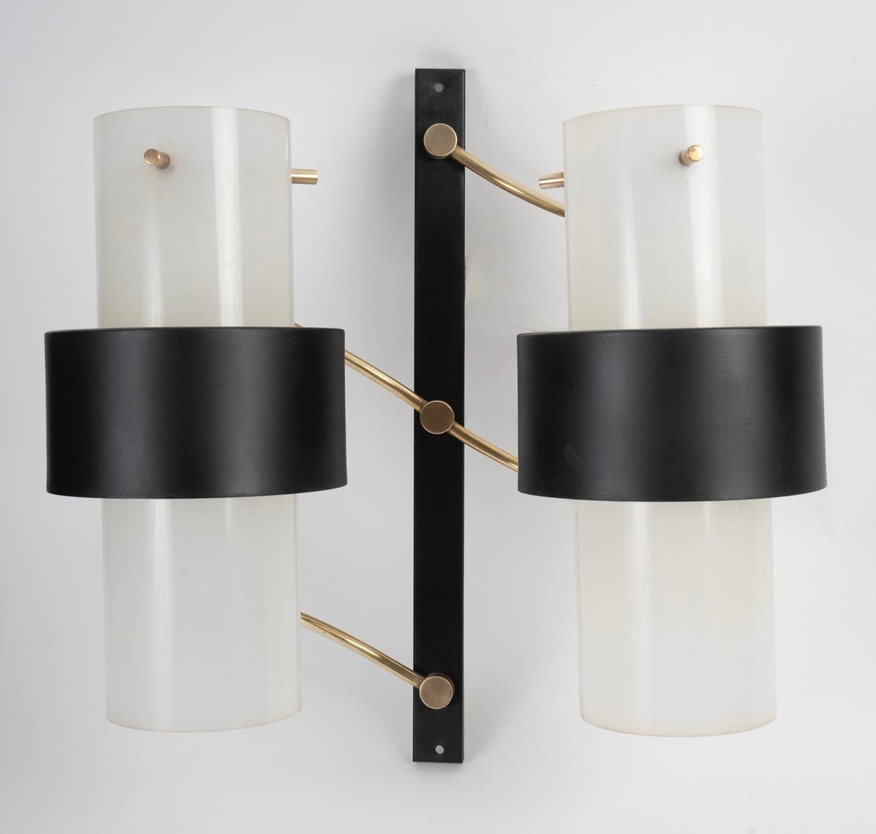 Composed of a vertically positioned black rectangular wall bracket on which rest 3 obliquely placed gilded brass rods, supporting two large perplex cylinders on which rest two black sheet metal cylinders positioned in the center of the sconce. 
They