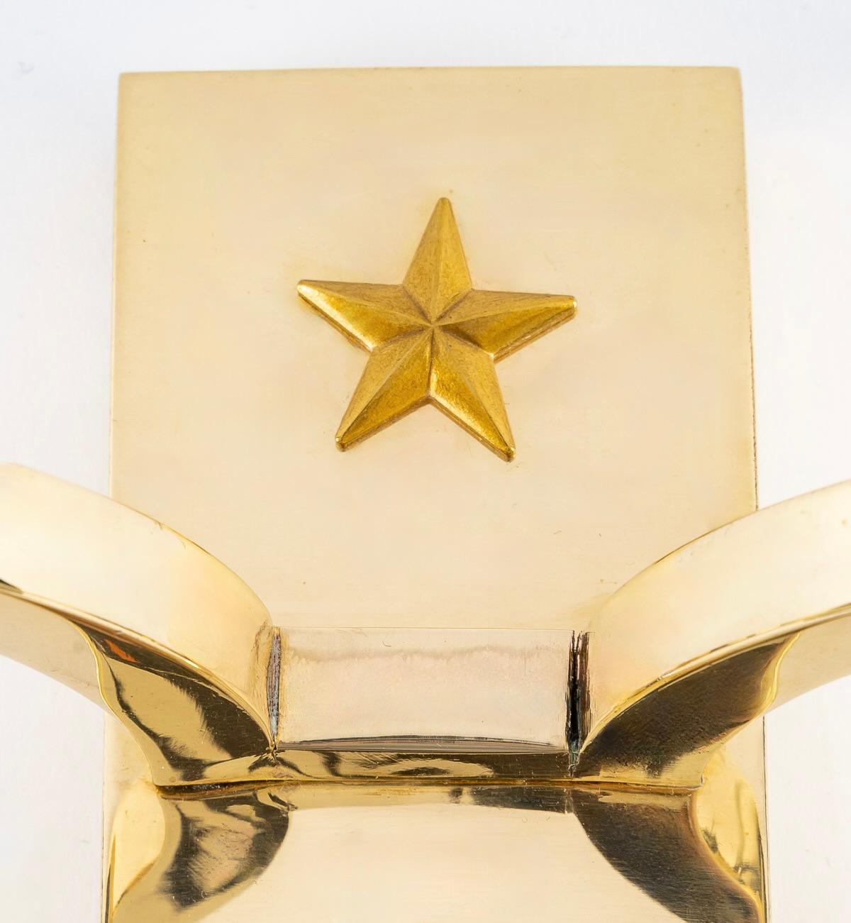 Composée d'une plaque murale en laiton doré de forme rectangulaire, elle est décorée de deux étoiles réparties sur le haut et le bas de l'applique.
Deux bras de lumière placés au centre de chaque côté de l'applique sont habillés d'abat-jour
