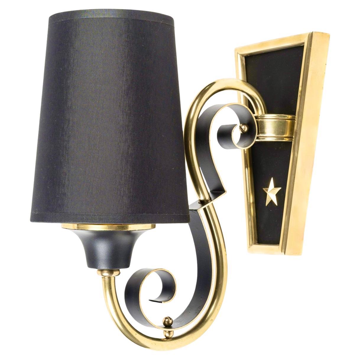 Comprising a trapezoidal wall plate in gilded and blackened brass, decorated with a gilded brass star, on which rests a round gilded brass rod acting as a light arm rising upwards, enhanced by a plate matching the shape of the wall light in