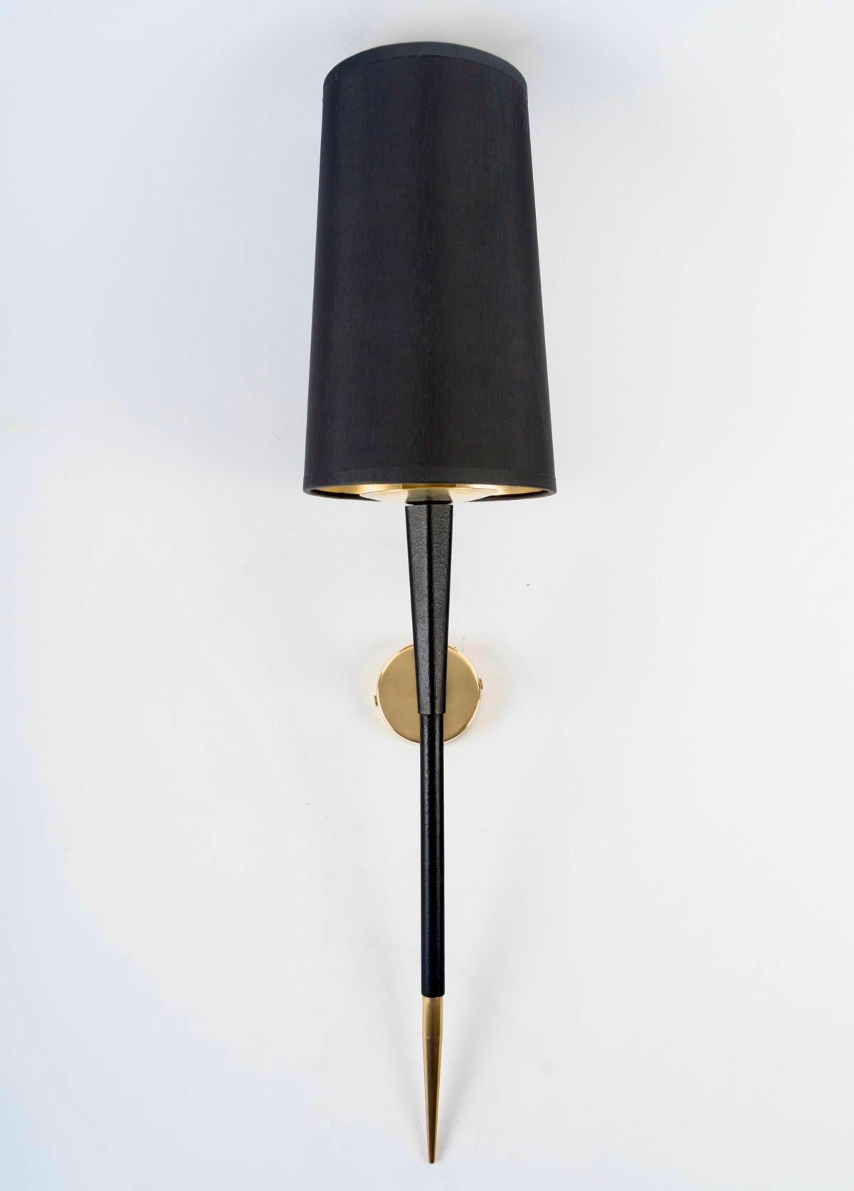 Composed of a round wall bracket in gilded brass on which rests the black wrought iron light arm, decorated with a gilded brass tip on the lower part.
On the upper part, it is dressed with a white cotton trapezoidal shade, highlighted at its base by