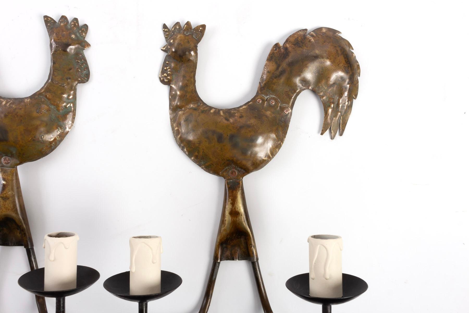 The upper part of the sconce features a rooster formed by hammered and set brass plates, hand-cut and given a brown patina. 
On the lower part, two upward-facing black wrought-iron luminous arms are underlined by cups and distributed on either side
