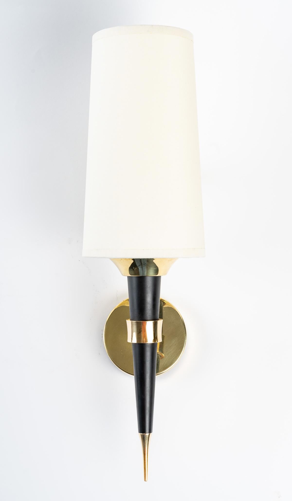 Composed of a blackened wooden rod decorated with a gilded brass tip on the lower part and a ring located in the middle of the arm, it is fixed to the round wall support by a rod, all in gilded brass.
An off-white cotton lampshade dresses the wall