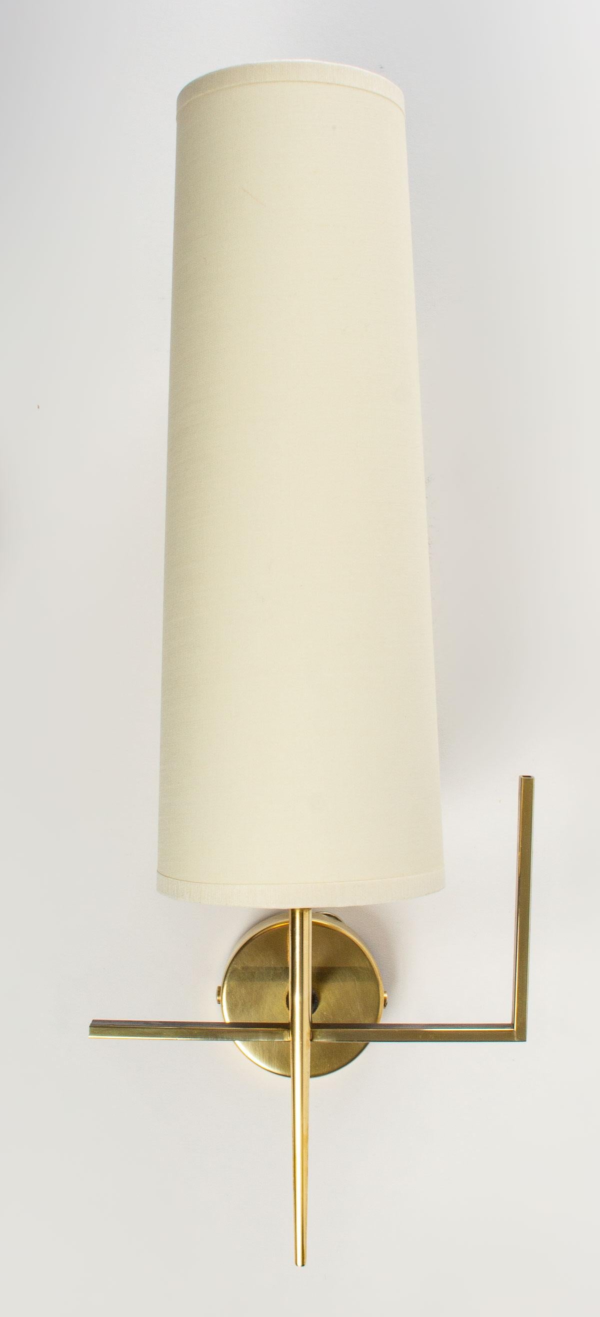 Composed of a round wall base, it serves as a support for the light arm placed vertically in the shape of a point, all in gilded brass. It is dressed with a trapezoidal shade of off-white color.
The sconce is decorated with a thin square section rod