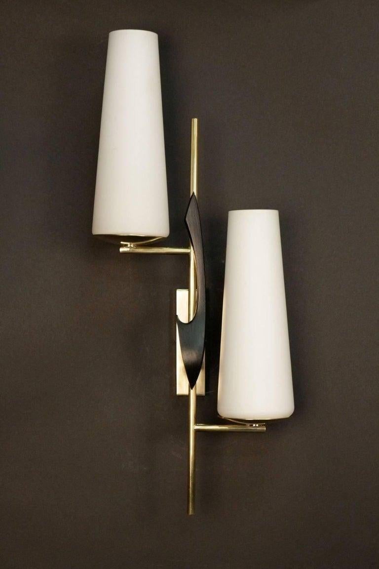 Der Wandsockel besteht aus einem Rechteck aus vergoldetem Messing und ist auf der Vorderseite mit einem Komma aus geschwärztem Messing verziert. Zwischen der Wandhalterung und der Dekoration befindet sich eine Stange, die zwei Lichtarme trägt, die