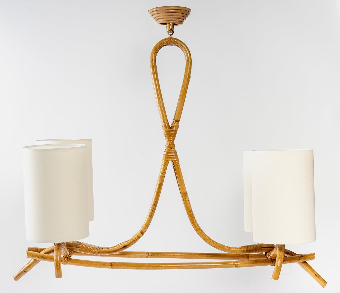 Composed of rattan rods laid horizontally on which rest at the ends four arms of light dressed with lampshades of cylindrical form in off-white cotton redone identically.
The whole is suspended by a rod in rattan posed vertically forming a loop on