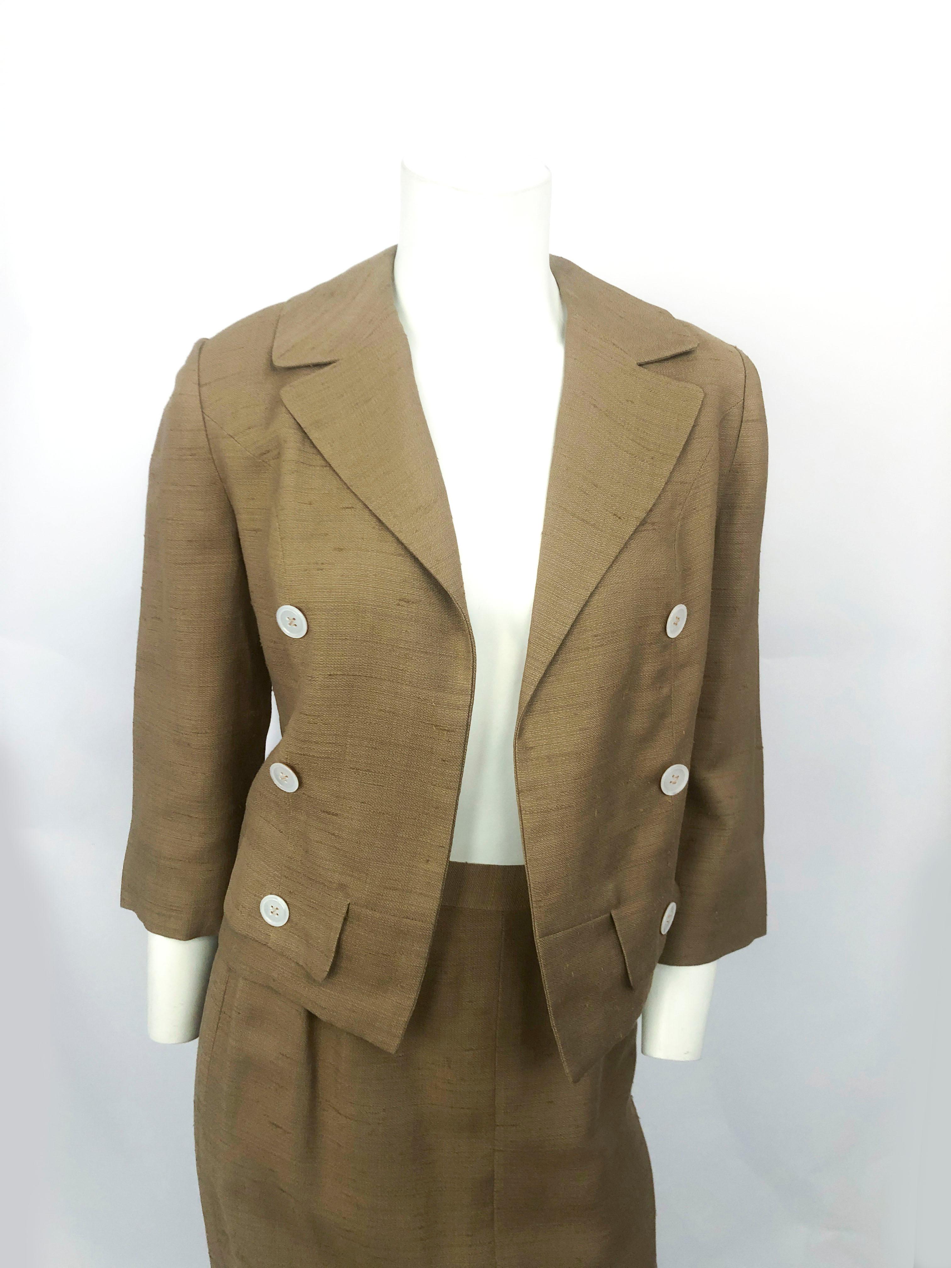 Late 1950s to early 1960s muted gold I. Magnin raw silk suit with abalone shell buttons, peak lapel collar, faux pockets, and glove-length sleeves. The jacket is bolero style. The skirt has a straight silhouette and has a zip closure along the side.