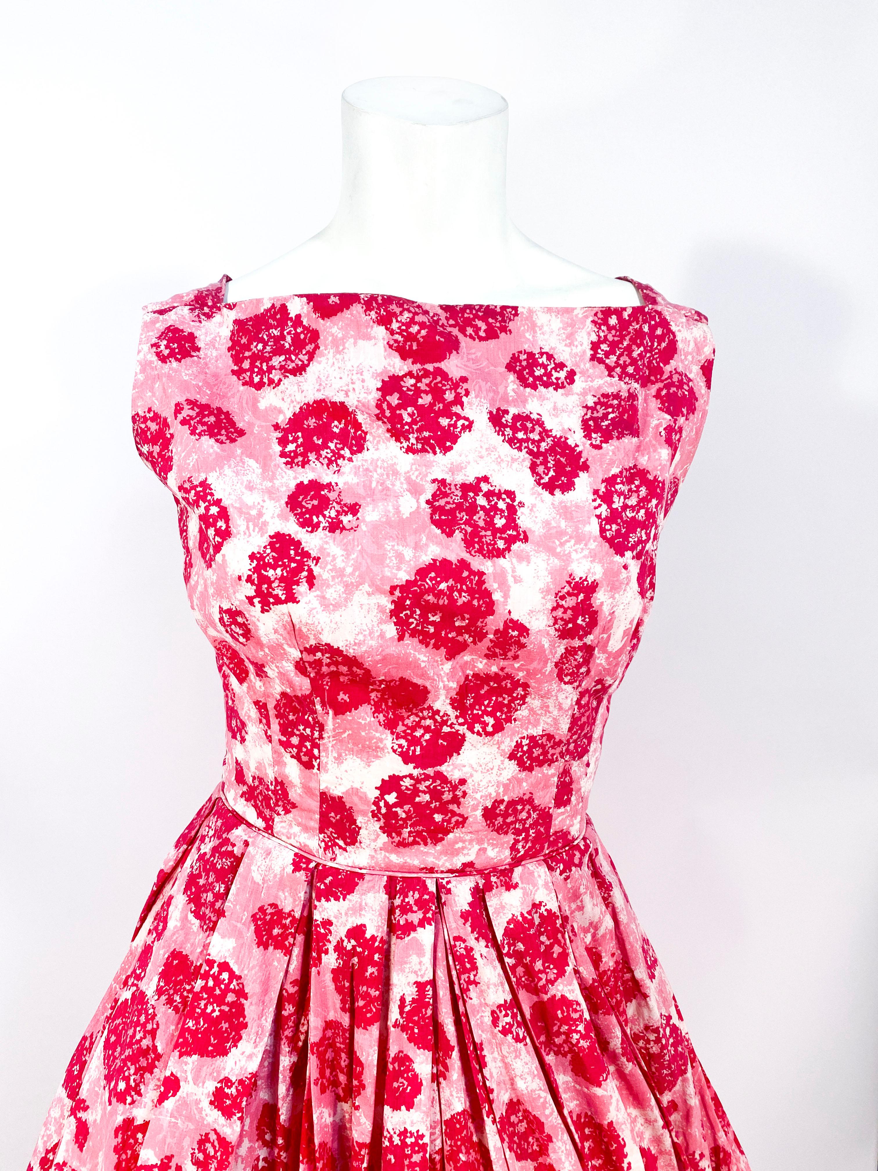 Late 1950s to early 1960s cotton day dress featuring an abstract floral print in several shades of pink. The structure of this dress has a full skirt, high neckline, and piped waist. The back of the dress has a gathered neckline, hidden zipper, and