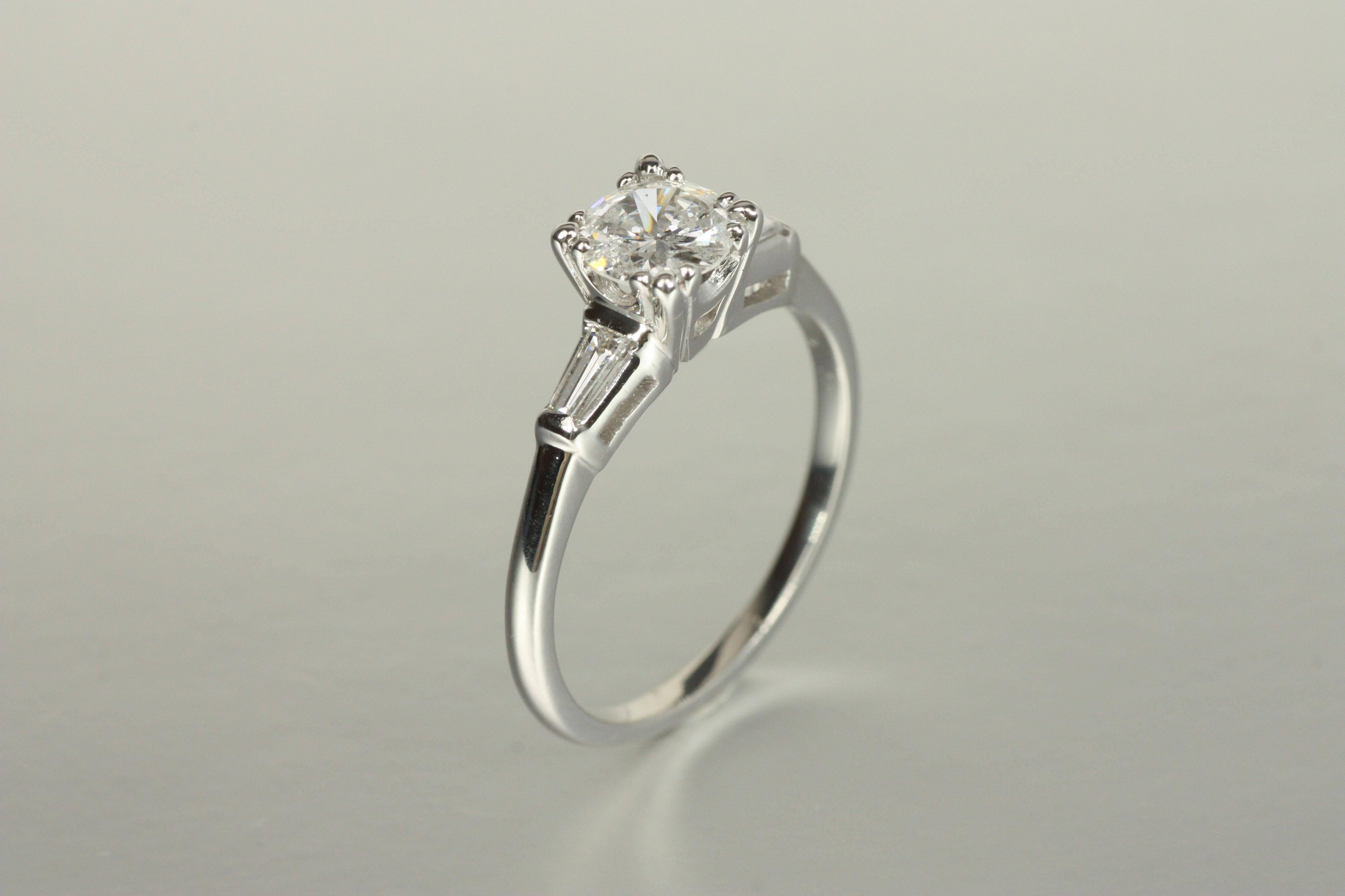 A timeless mid-century classic, this vintage 1950s 3-stone engagement ring in platinum features a 0.64ct round brilliant cut diamond at its center, framed by tapered baguette diamonds for a sleek, refined look. A perfect engagement ring for lovers