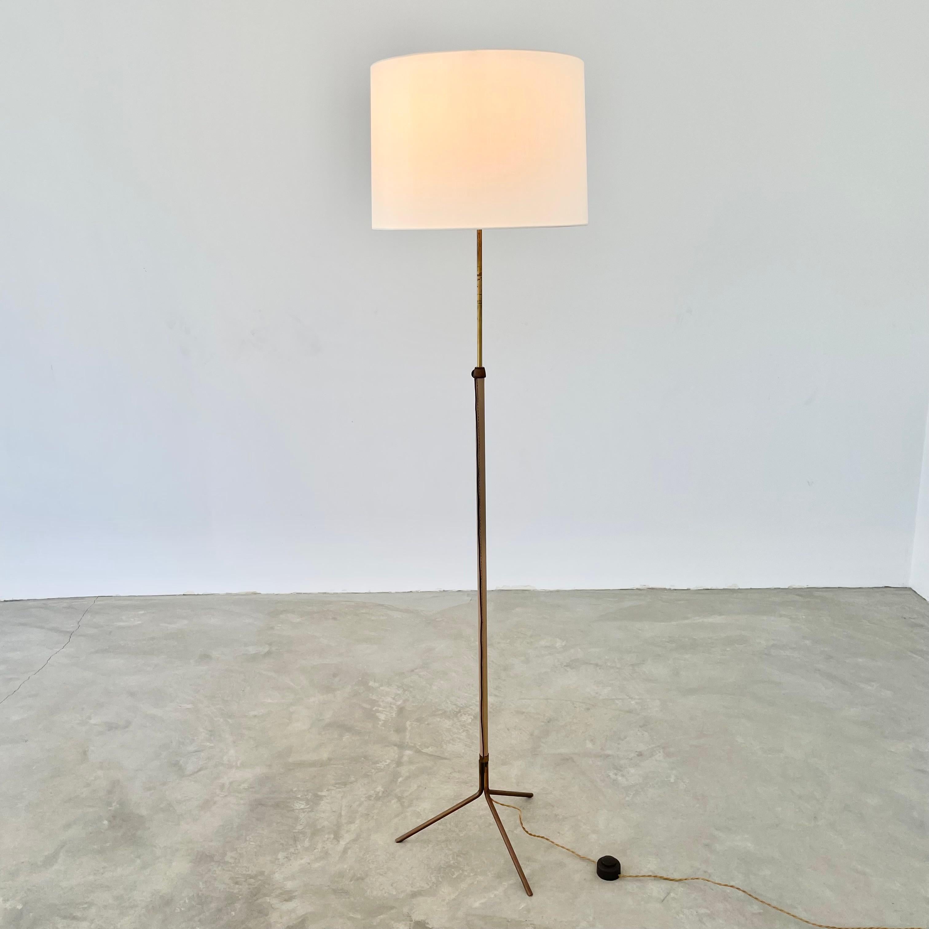 Handsome brass and leather floor lamp in the style of French designer Jacques Adnet. Vers les années 1950. Beige leather wrapping the full length of lamp. Height adjustable brass rod runs inside. Signature Adnet contrast stitching. Lamp has been