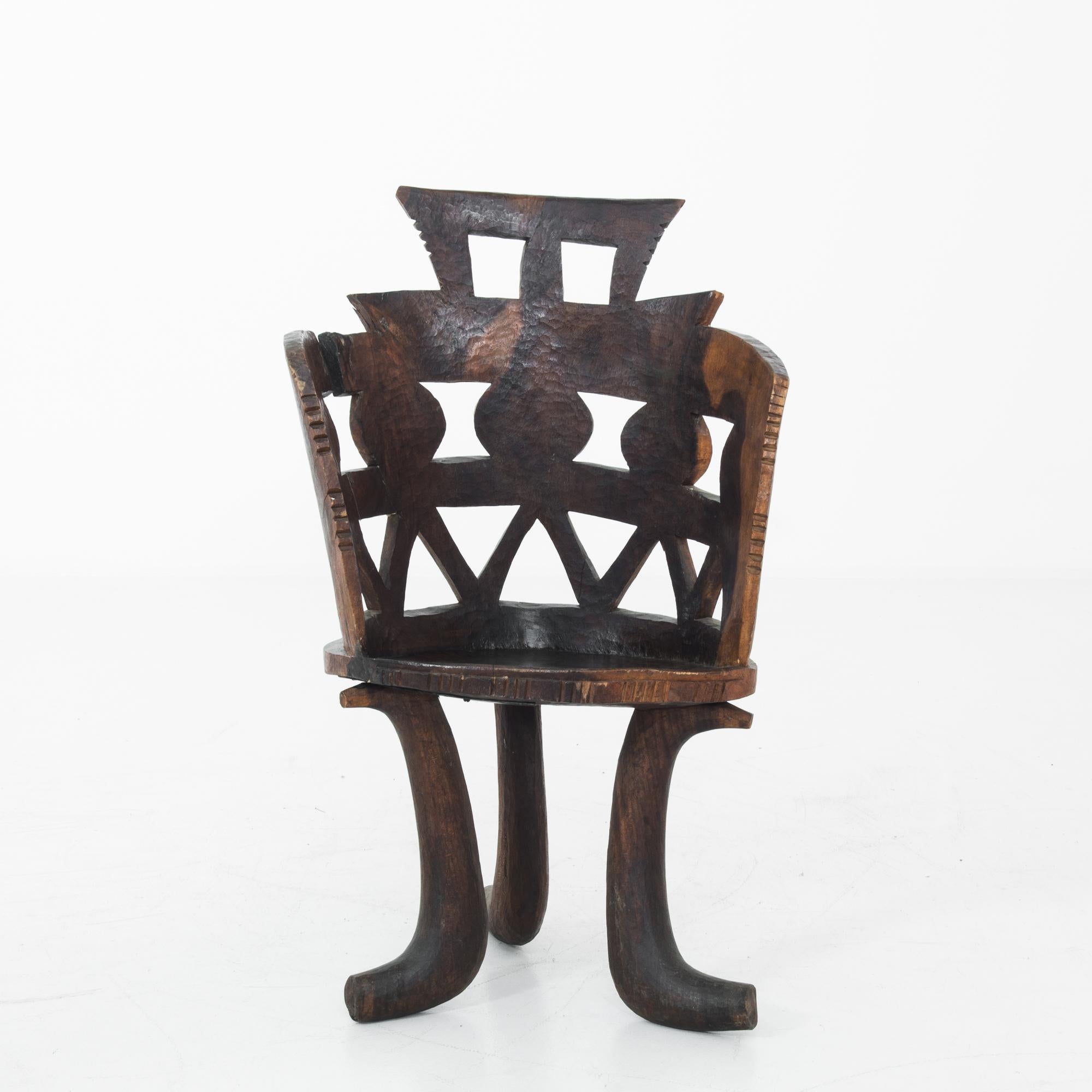 A wooden armchair from the African continent, produced circa 1950. Things may fall apart, but this hand carved chair was built to last. Standing on three legs that curve like elephant tusks, the seat back is an image of unity: depicting people