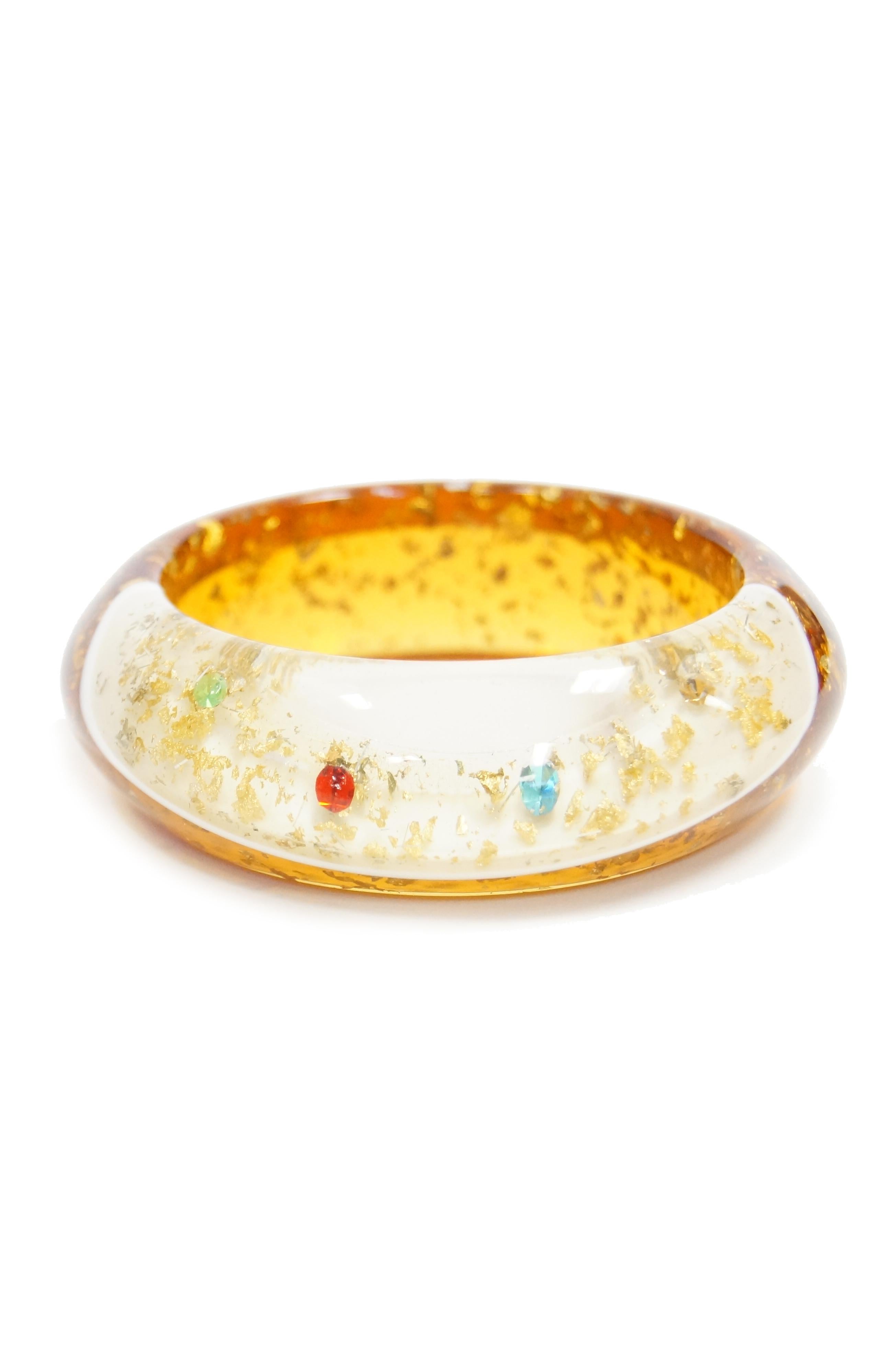 Playful wide amber lucite bangle! This bangle is primarily an orange - gold amber color and also a slanted band of opaque white as well as a pocket of clear lucite. The bracelet is adorned with shimmering gold glitter, and features three blue, red,