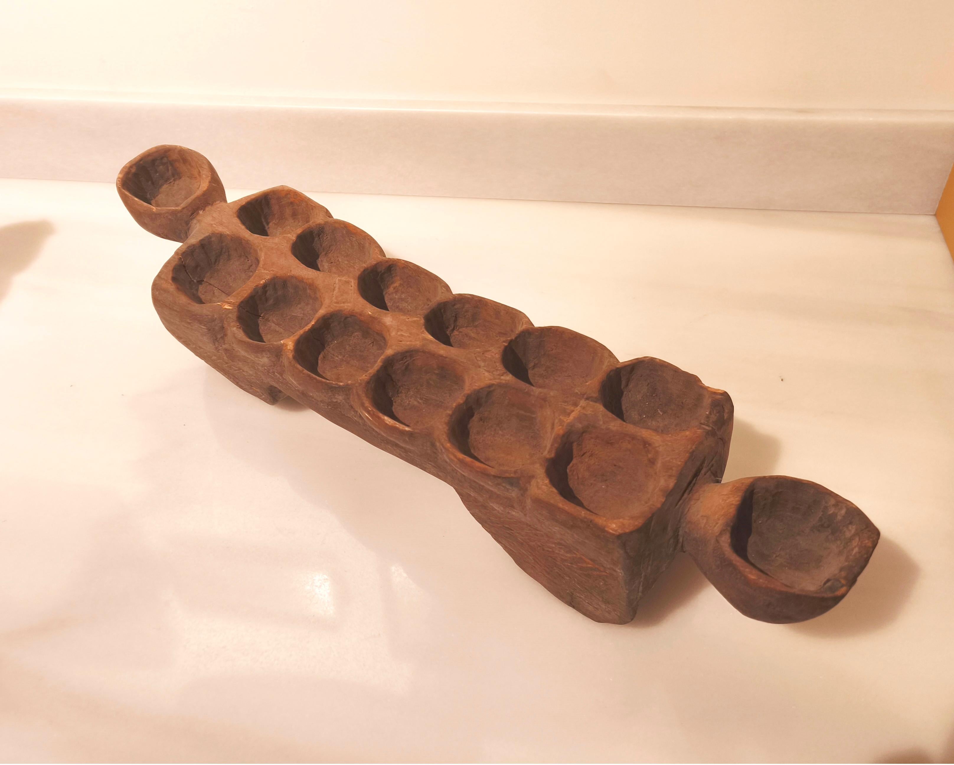 Pièce artisanale sculptée dans du bois massif utilisée pour le jeu traditionnel africain du Mancala. Il présente douze cavités centrales et deux plus grandes aux extrémités, typiques de ce jeu de stratégie et de collecte de graines. Sa forme