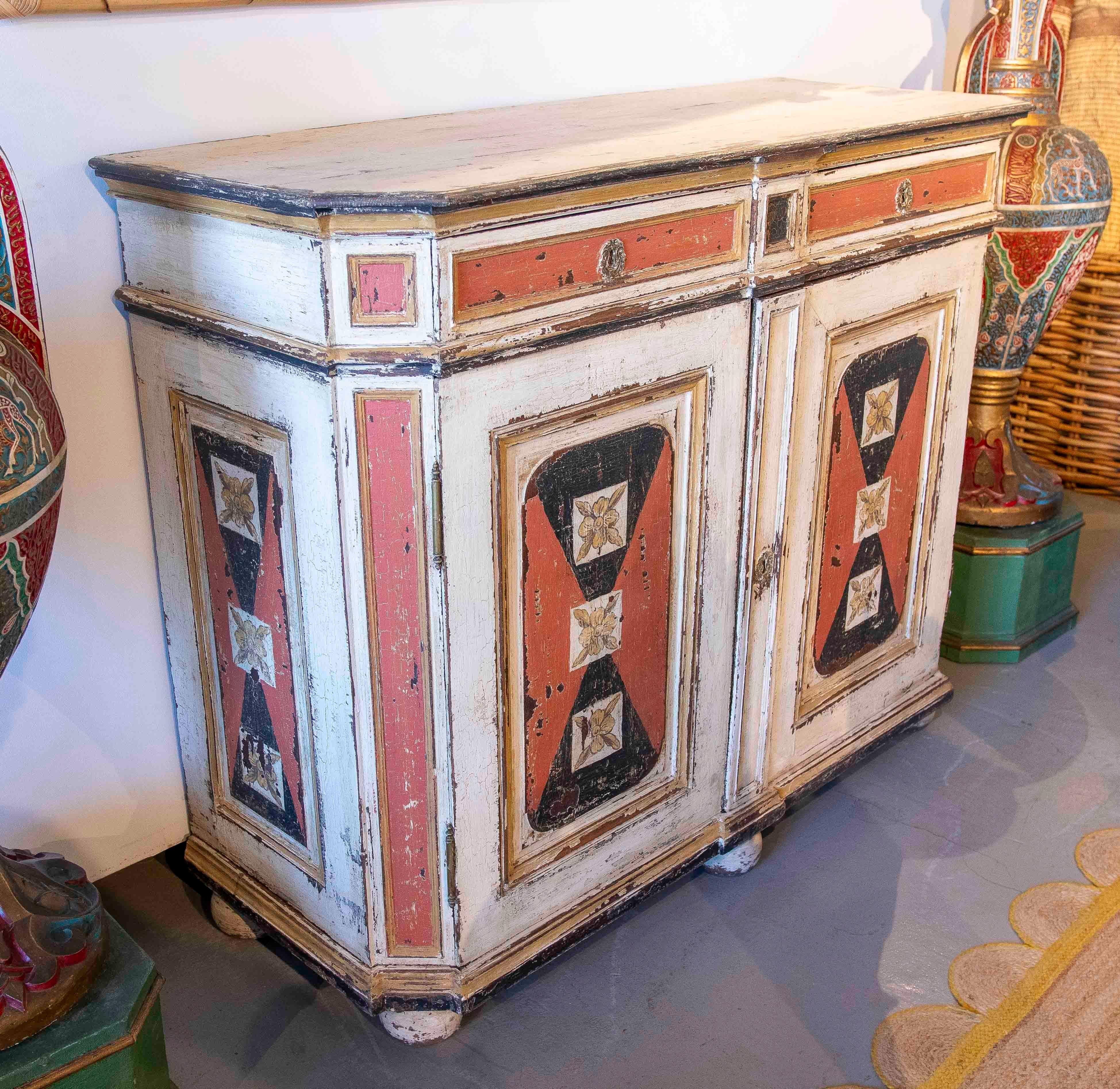 Buffet ancien à deux portes, au cadre robuste et à la finition vieillie, décoré de panneaux peints à la main en rouge terracotta, noir et crème. Chaque porte présente un motif symétrique de triangles opposés encadrant des fleurs stylisées, créant un