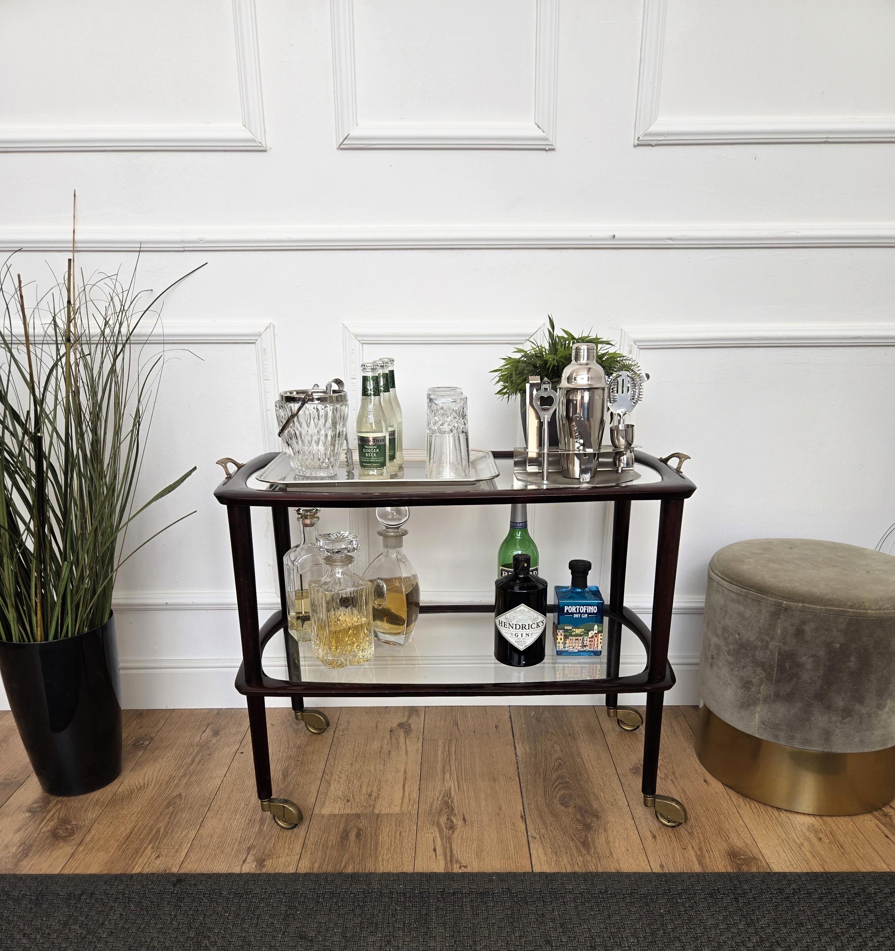 Eleganza e raffinatezza del carrello bar a secco in stile Art Déco con due ripiani in vetro, evidenziate dal grande design della struttura in legno e dalla forma complessiva completata dai dettagli delle ruote e delle maniglie originali in ottone