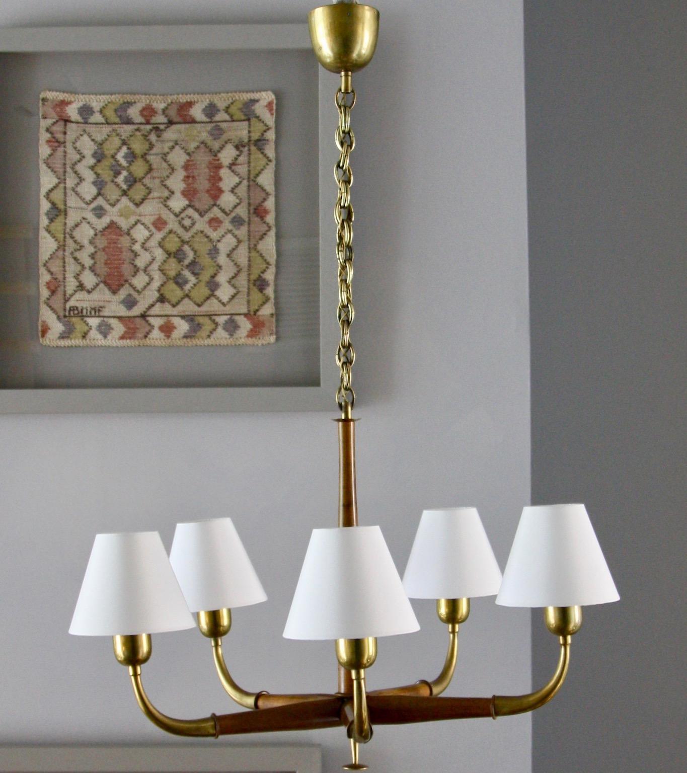 An distinctive pendant light featuring carved mahogany sections, brass details and neutral candle lampshades, Austria, circa 1940-1955.
The interplay between the brass and mahogany that make-up the arms of the chandelier is aesthetically very