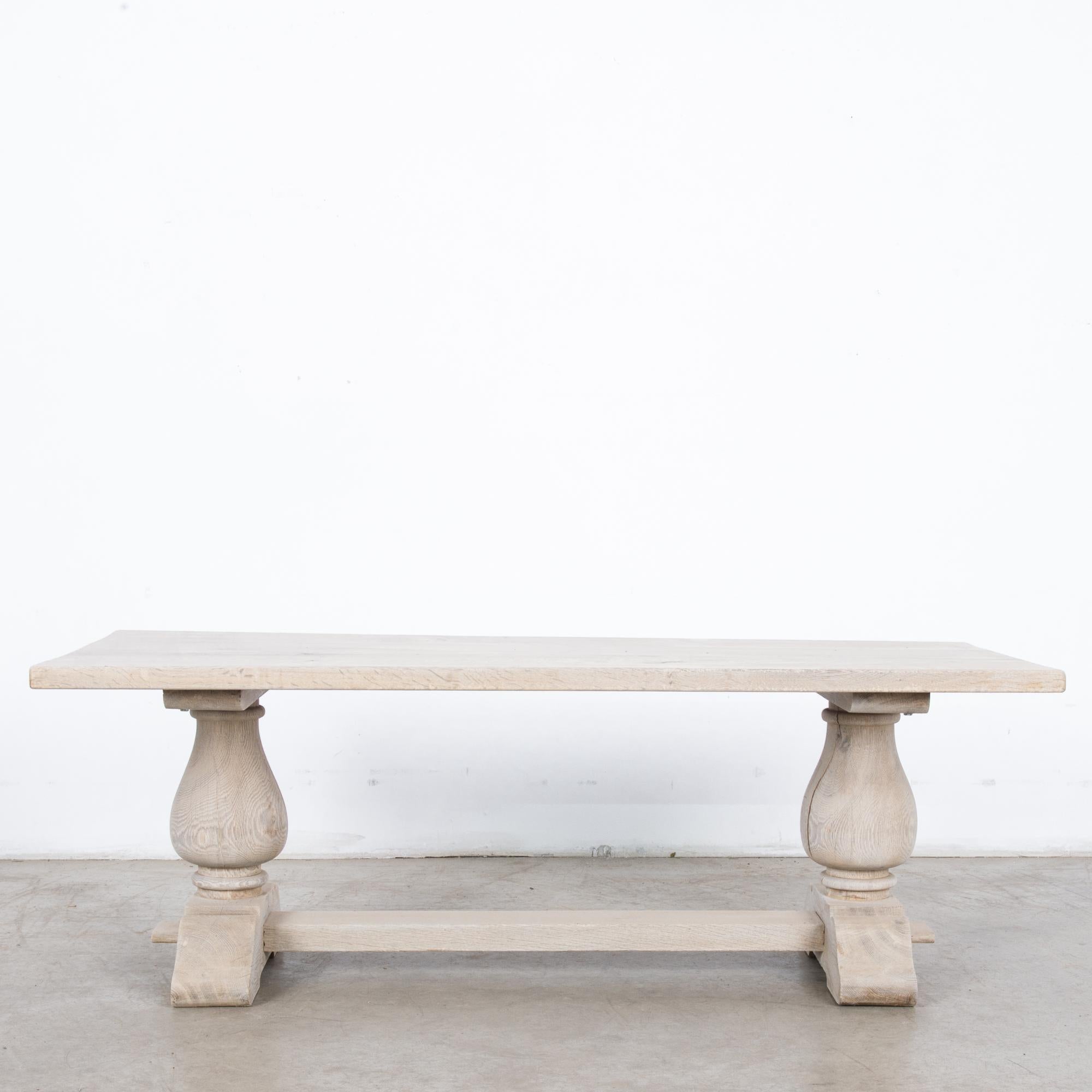A bleached oak table from Belgium, circa 1950. The smooth rectangular tabletop sits upon a pair of trestle legs, which bloom into broad tulip shapes. A strut joins the brackets of the feet — broad and sturdy, with an elegant profile. The oak has