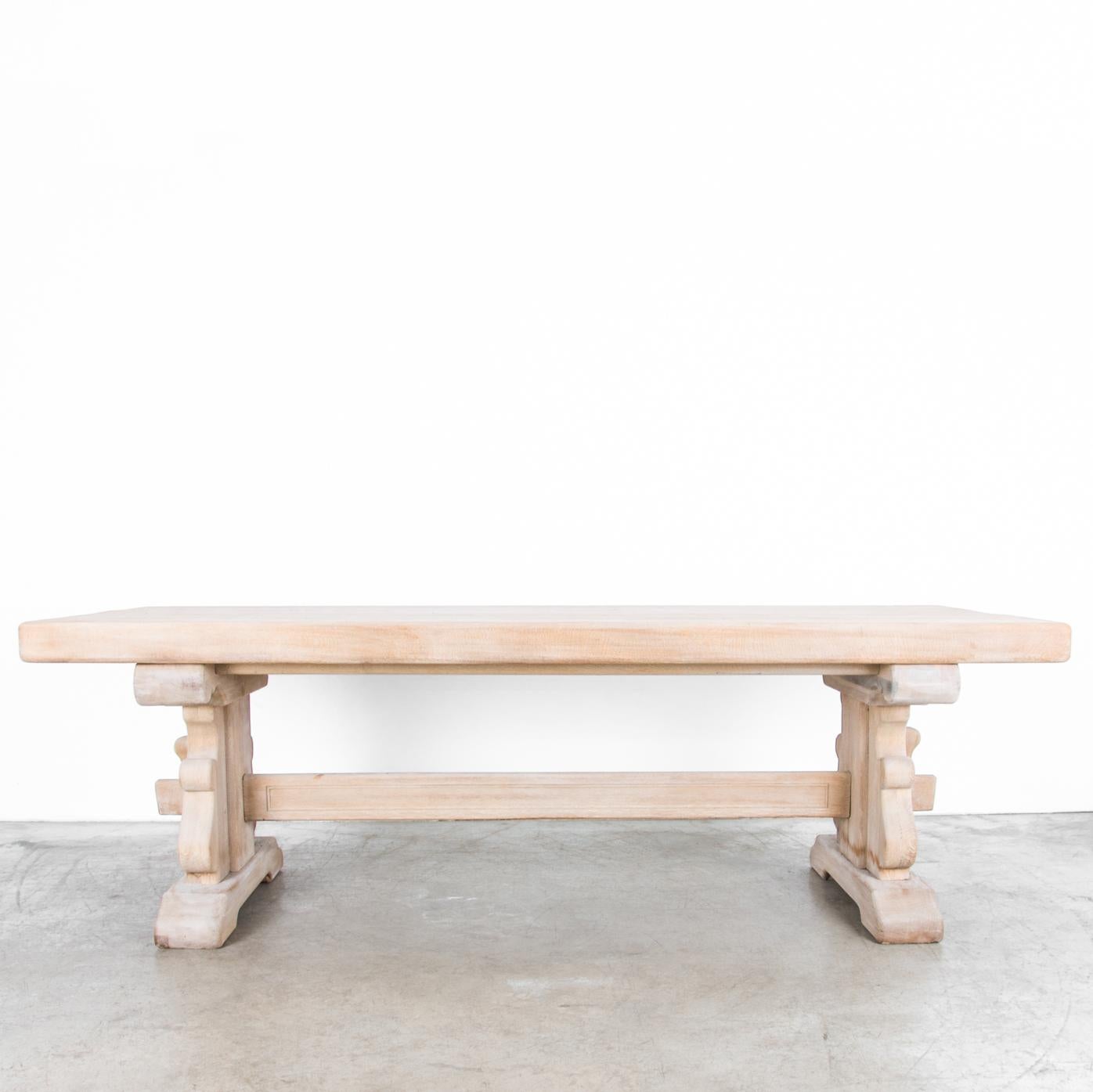 A bleached oak dining table made in Belgium, circa 1950, featuring trestle legs with a curved, heraldic profile. The legs are joined by a strut held in place with pegs and slats, yielding a design that is both stately and provincial. The sturdy