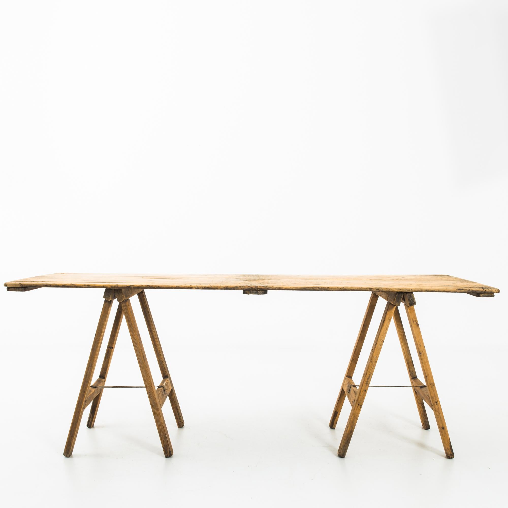 A wooden table from 1950s Belgium. A light tabletop of wooden boards rests atop a pair of A-frame legs, creating an industrial aesthetic. The wood has a warm, honeyed tone and a rustic finish; a light patina clouds the boards of the tabletop.