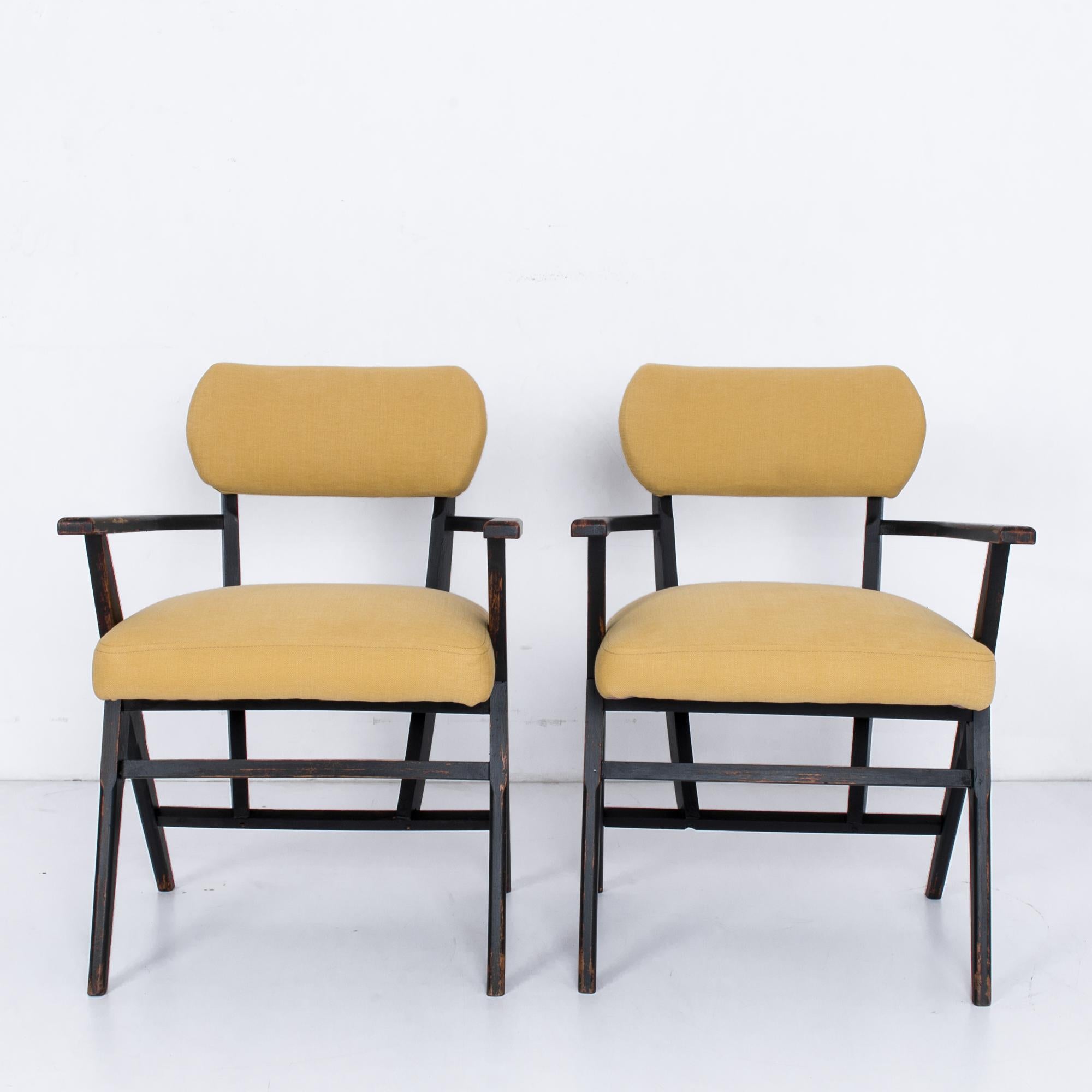 A pair of 1950s armchairs from Belgium, with a wooden frame and upholstered seat and backrest. The light golden yellow fabric makes a stylish contrast with the dark burnished wood. The design mimics the casual shape of a folding chair, with a