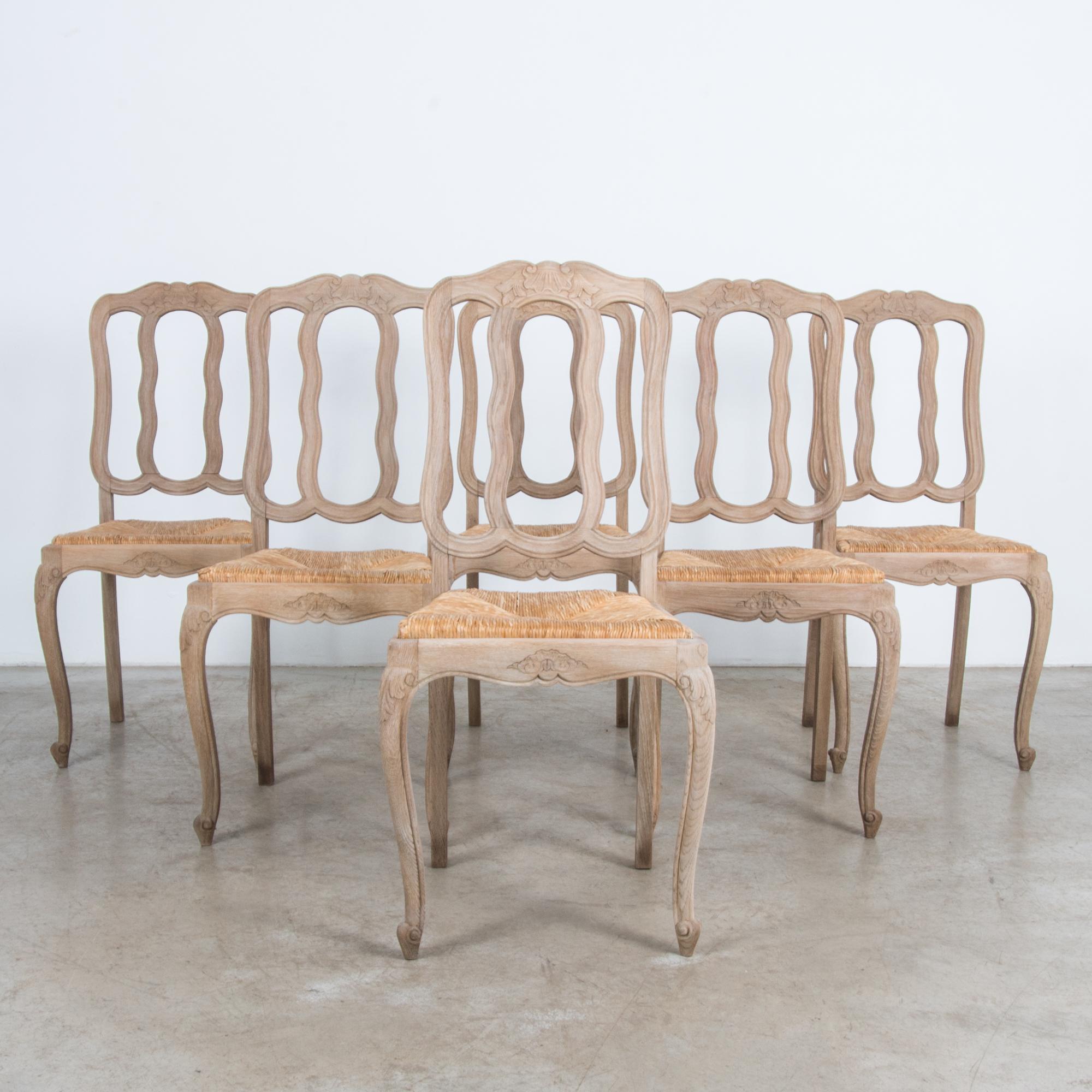 This set of six oak dining chairs from Belgium, circa 1950, combines a classical French shape with a wicker seat for a refined yet casual effect. The distinctive flowing curves of the chair back create soft loops that are echoed by scallop and