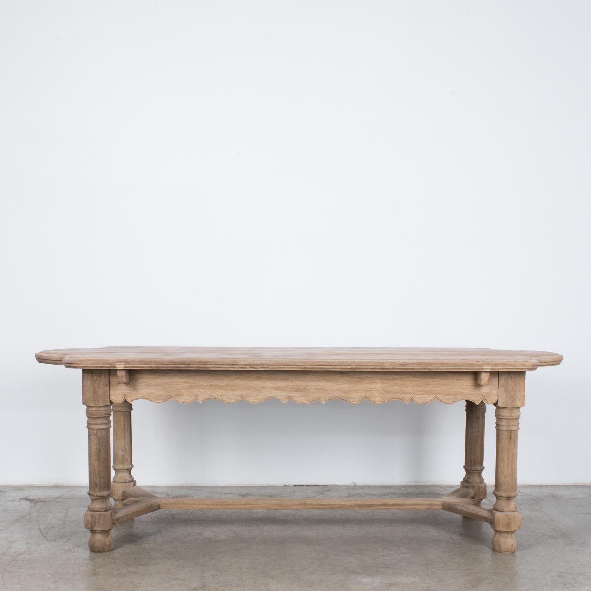 An oak dining table from Belgium, circa 1950. Robust turned legs support an elegantly contoured tabletop. The apron has a dramatic, scalloped border; the legs are joined by a sophisticated strut. The soft, blond color of the restored oak elevates