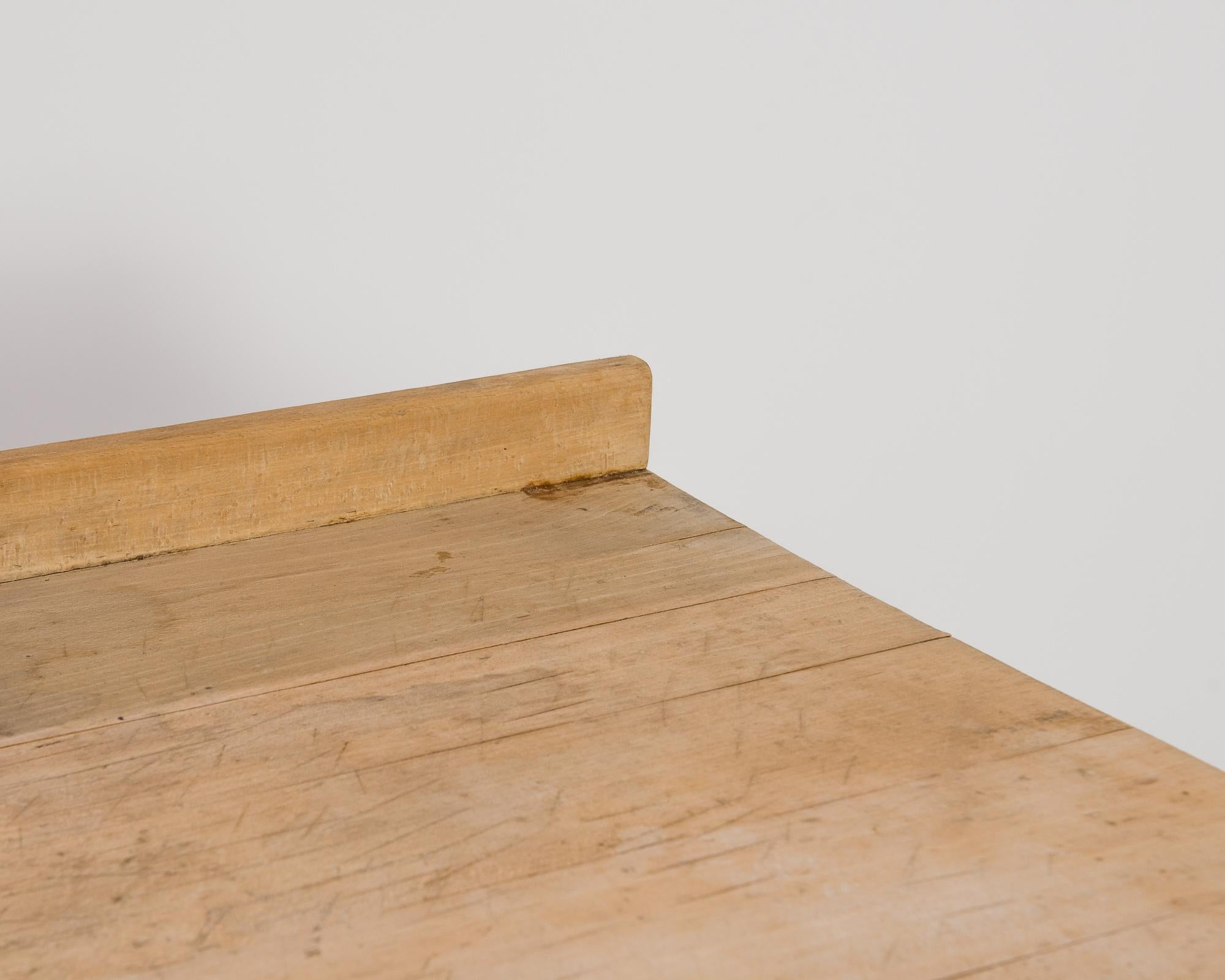 A wooden bakery table from Belgium, produced circa 1950. A wide, unadorned working surface takes center stage in this vintage bakery table. Featuring a raised lip along the back edge and an open base connected by box stretchers, this sturdy table