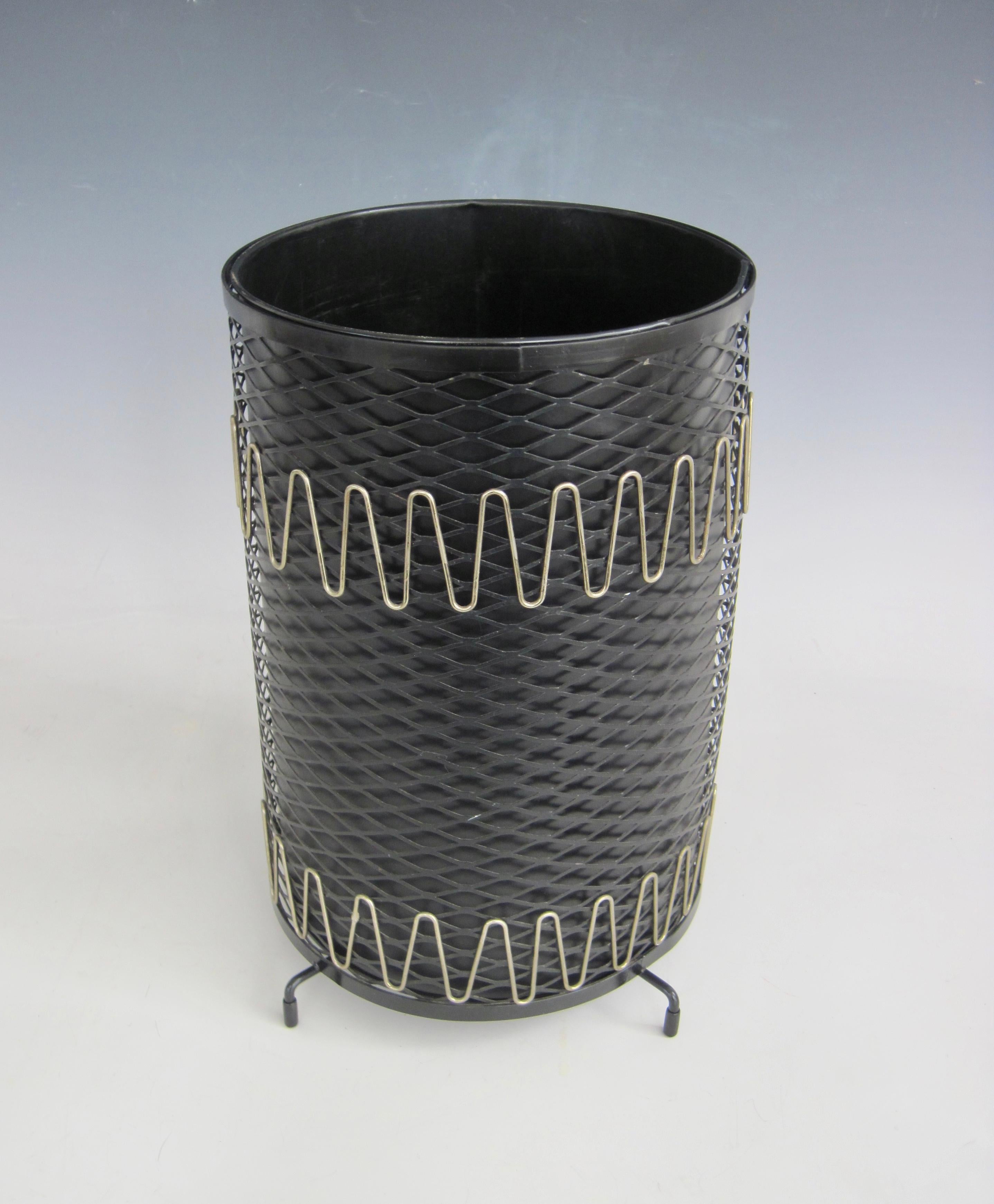 1950s atomic style black and brass wastebasket/trash receptacle. The wrought iron mesh outer sleeve has brass wave design near top and bottom edge, and  four-footed bottom. The aluminum inner sleeve can be removed to empty trash. 
