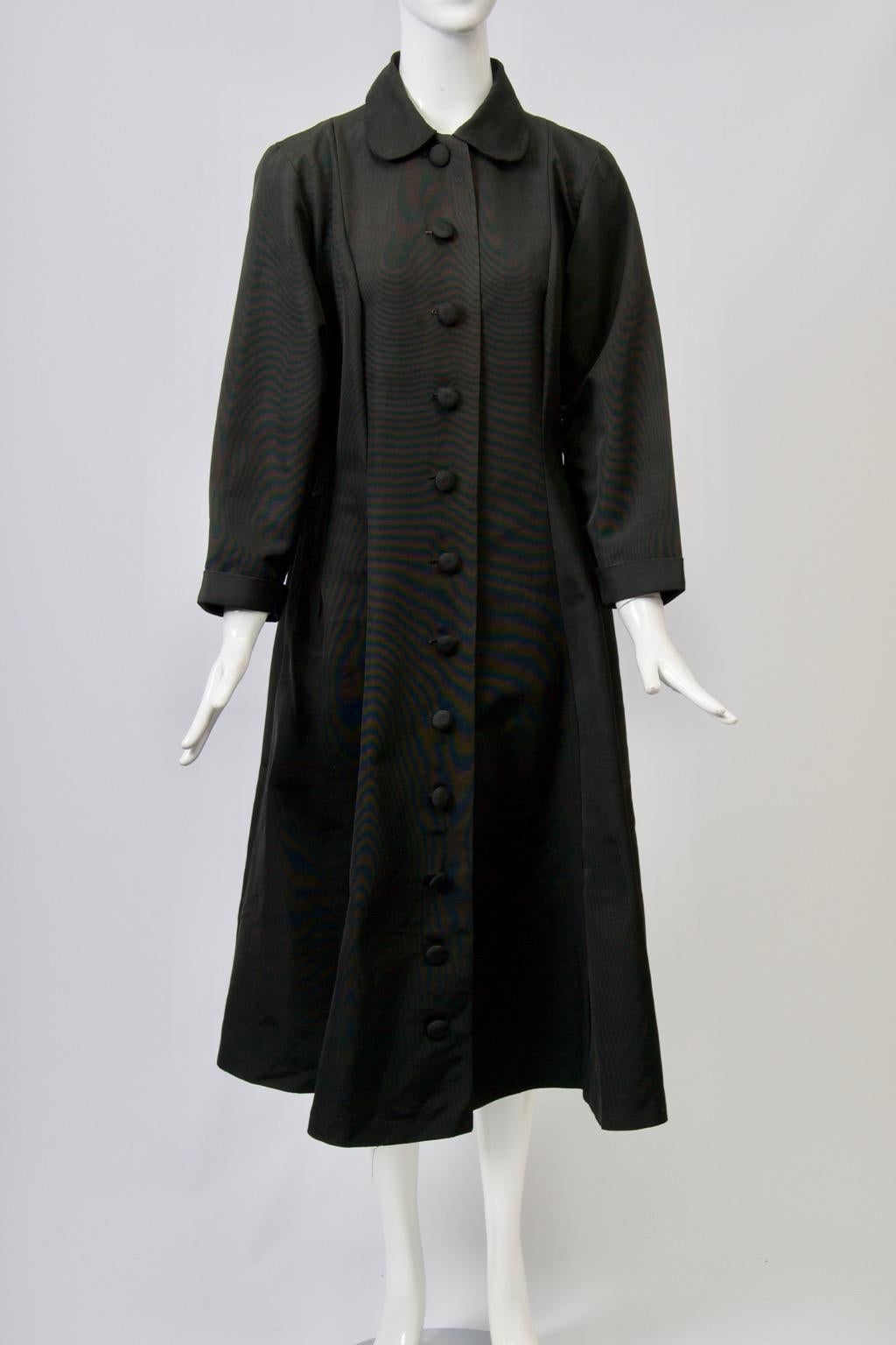 Schwarzer Vintage-Mantel aus Faille im Prinzessinnen-Stil mit Petermännchen-Kragen und selbstklebenden Knöpfen vom Hals bis zum Saum. Eingesetzte Ärmel mit tiefen Armlöchern. Vertikale Nähte formen die taillierte Taille und den ausgestellten Rock.