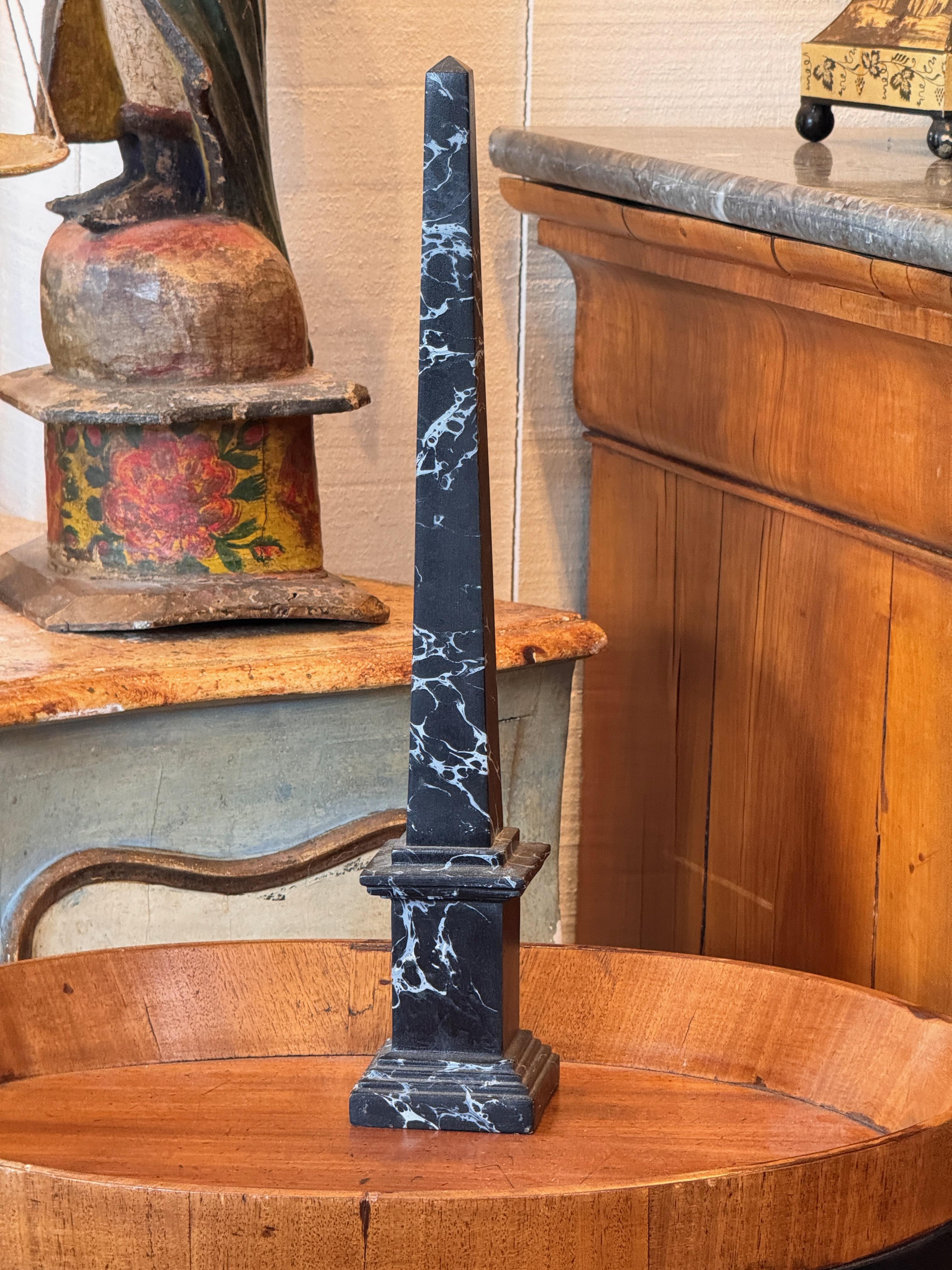 Alto obelisco decorativo realizzato in materiale composito nero con venature bianche in stile marmo. Posizionato su una base a gradini, con una silhouette pulita e angolare.