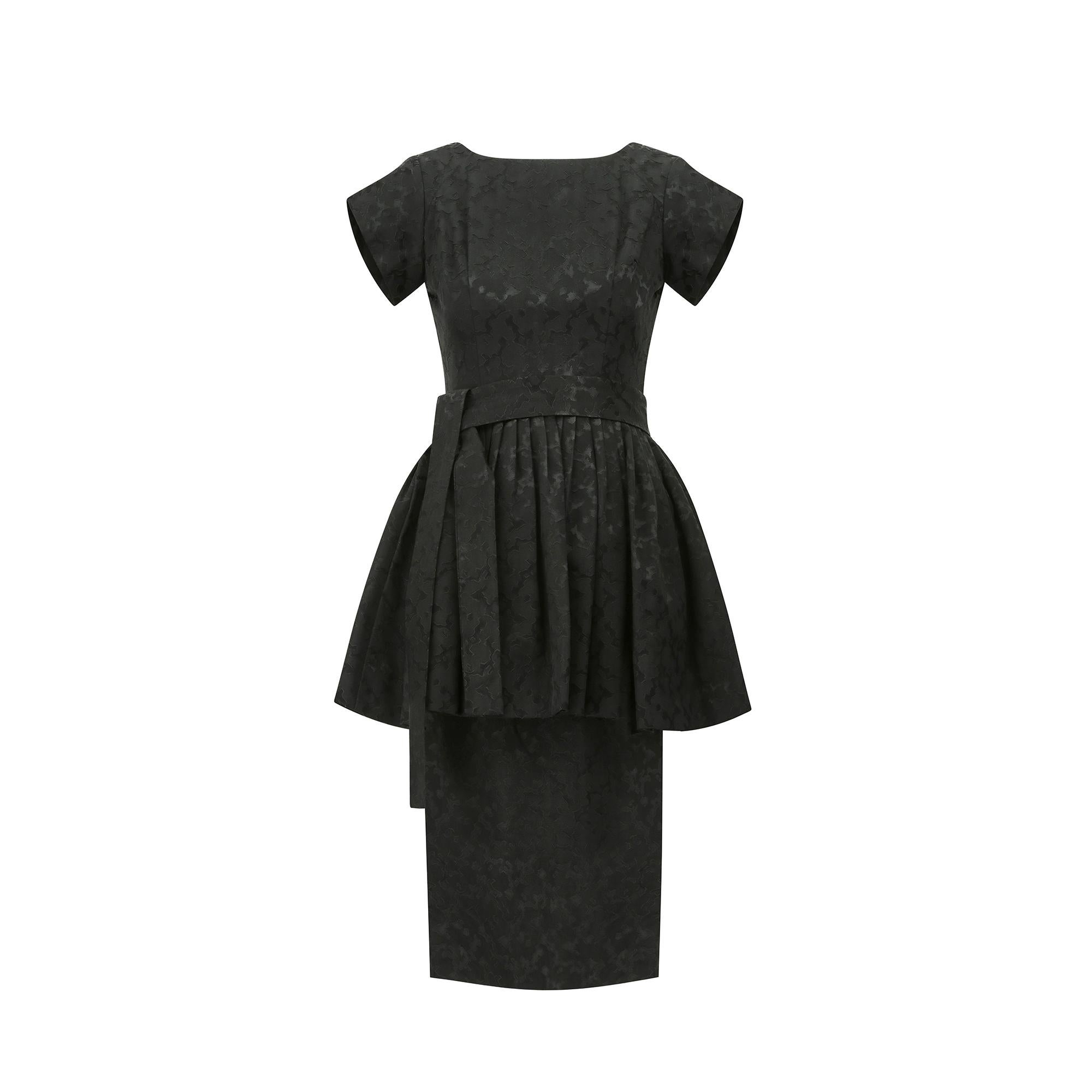 A timeless 1950s shape, this short-sleeve little black dress has a pencil skirt and a cinched waist and tie belt to accentuate the female figure. The voluminous peplum overskirt is crafted from the same fabric and is lined with layers of tulle for