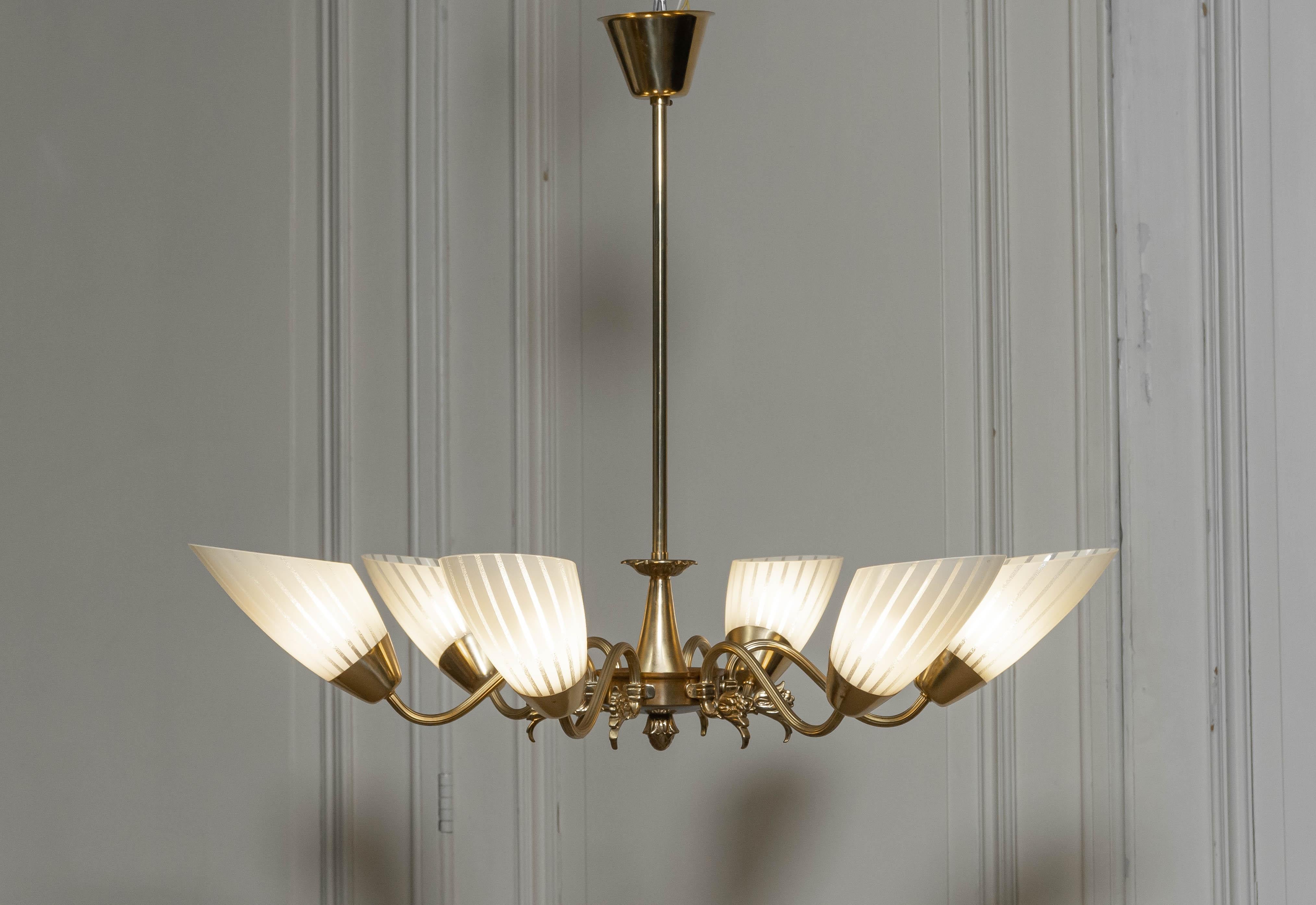 Il s'agit d'un lustre suédois en laiton des années 1950, doté de six bras élégamment courbés et ornés de décorations florales. Chaque bras supporte un abat-jour en verre d'art finement travaillé avec un motif rayé subtil, ajoutant une touche