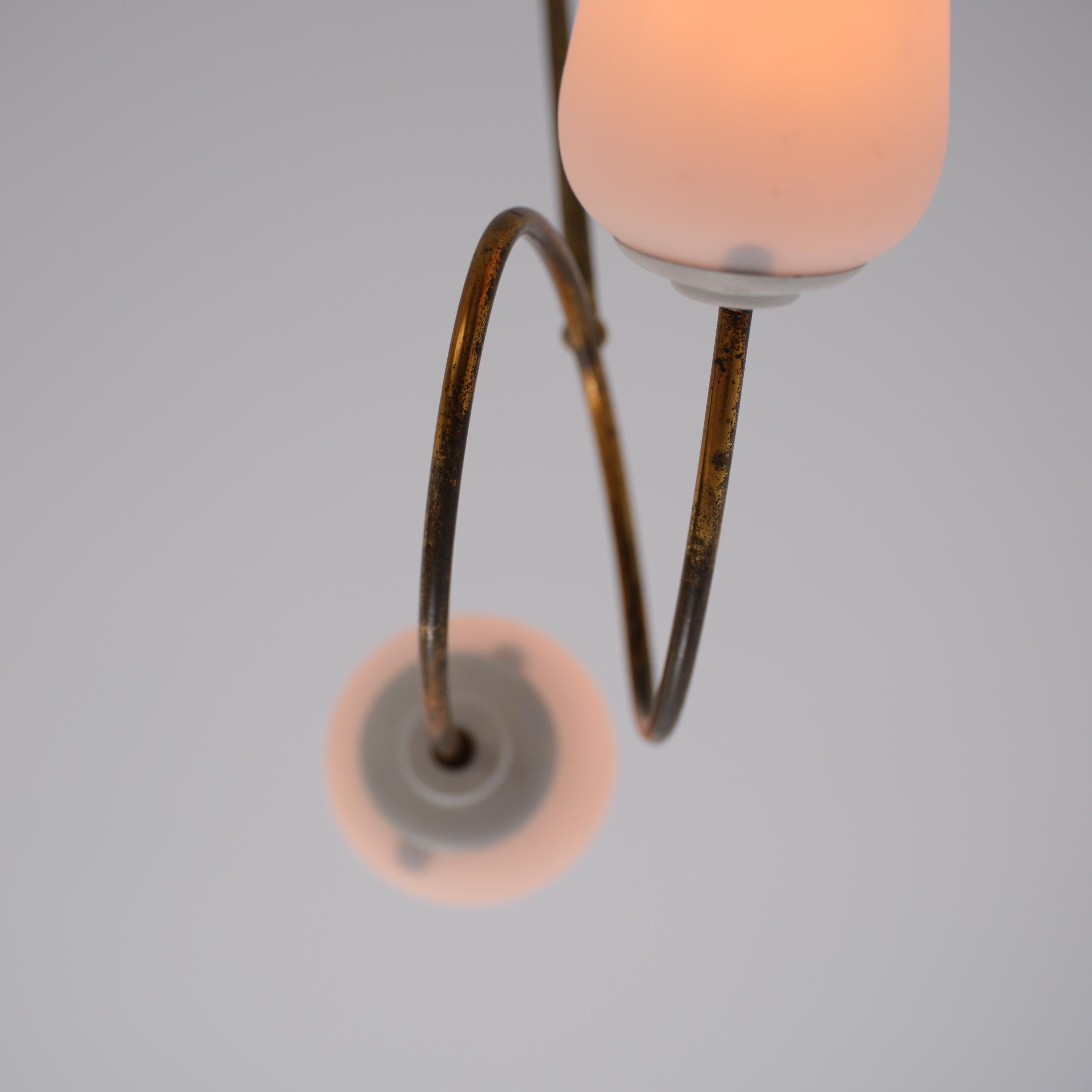 Lampadario a due luci degli anni '50 con stelo in ottone e anello centrale circolare che sostiene una coppia di bracci contrapposti. Ogni braccio sostiene un diffusore in vetro opalino angolato con un profilo morbido e affusolato. L'ottone originale