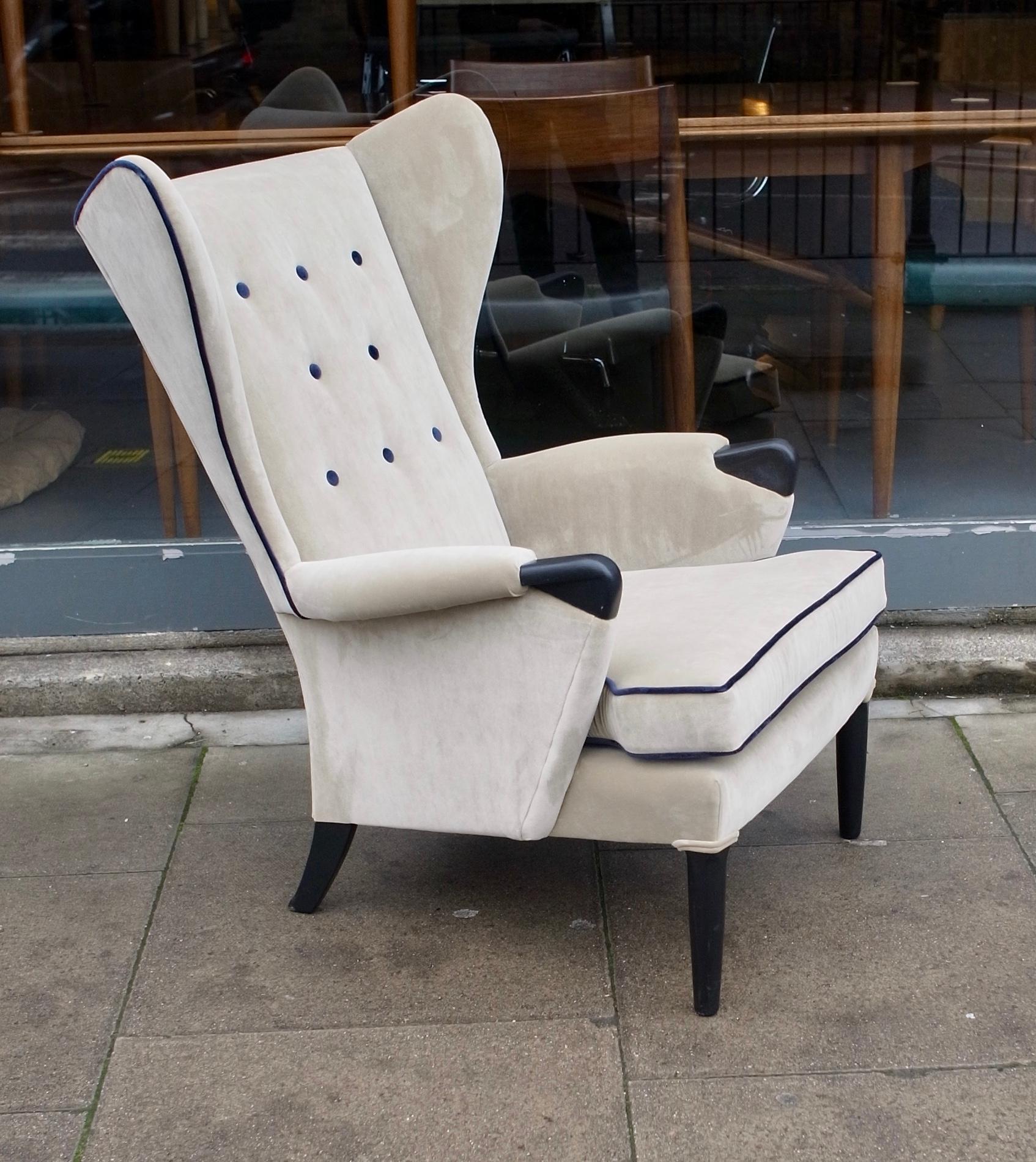A very comfortable and stylish vintage 1950s English buttoned wingback armchair. Newly reupholstered in an oatmeal coloured velvet textile, with navy velvet piping and buttons, that accentuates the chairs elegant profile. The black painted Beechwood
