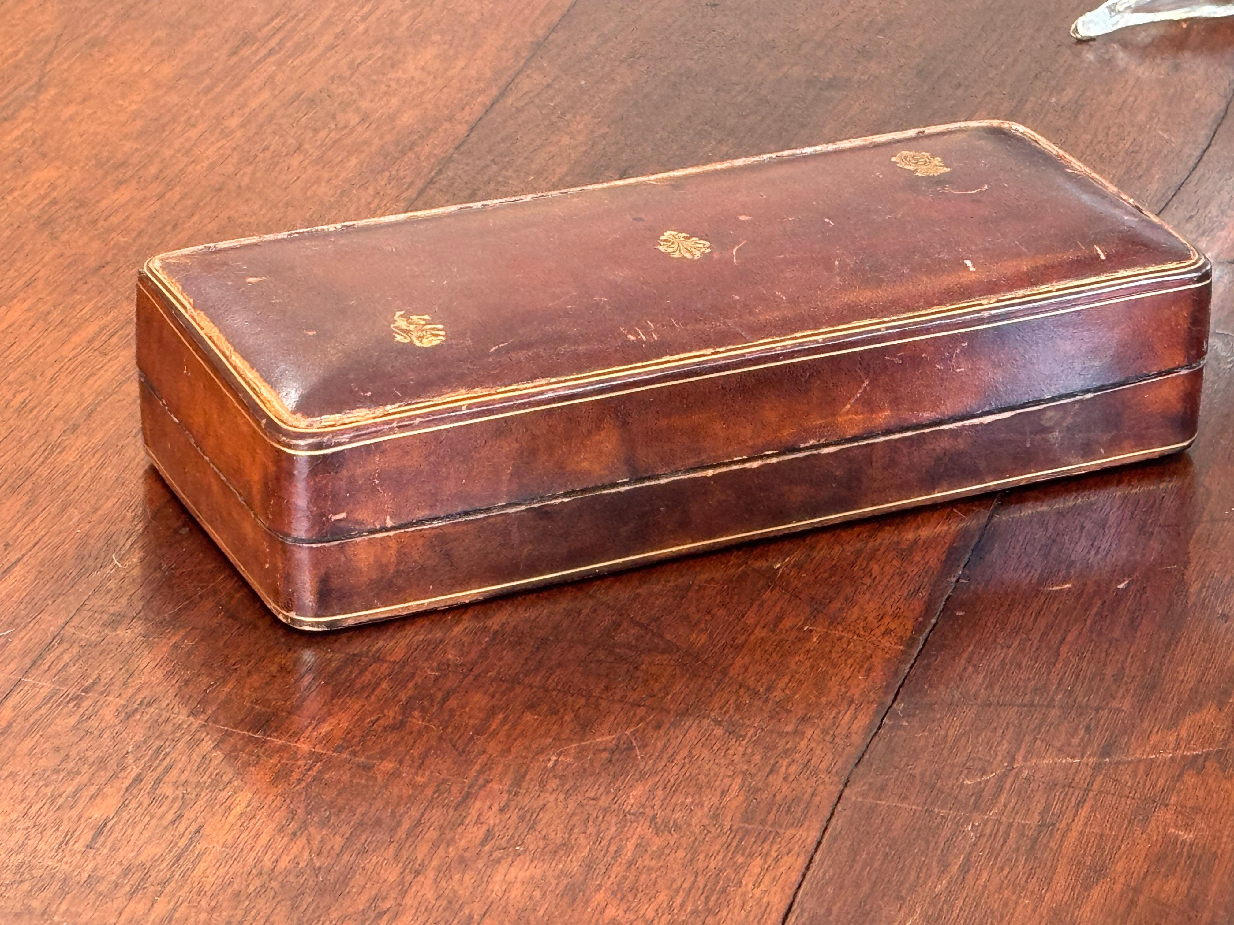 Una scatola rettangolare in pelle marrone con stemmi dorati sul coperchio e una sottile linea di bordo in oro. L'interno è foderato in pelle scamosciata con un unico scomparto. Un bel pezzo con una patina calda e proporzioni classiche.