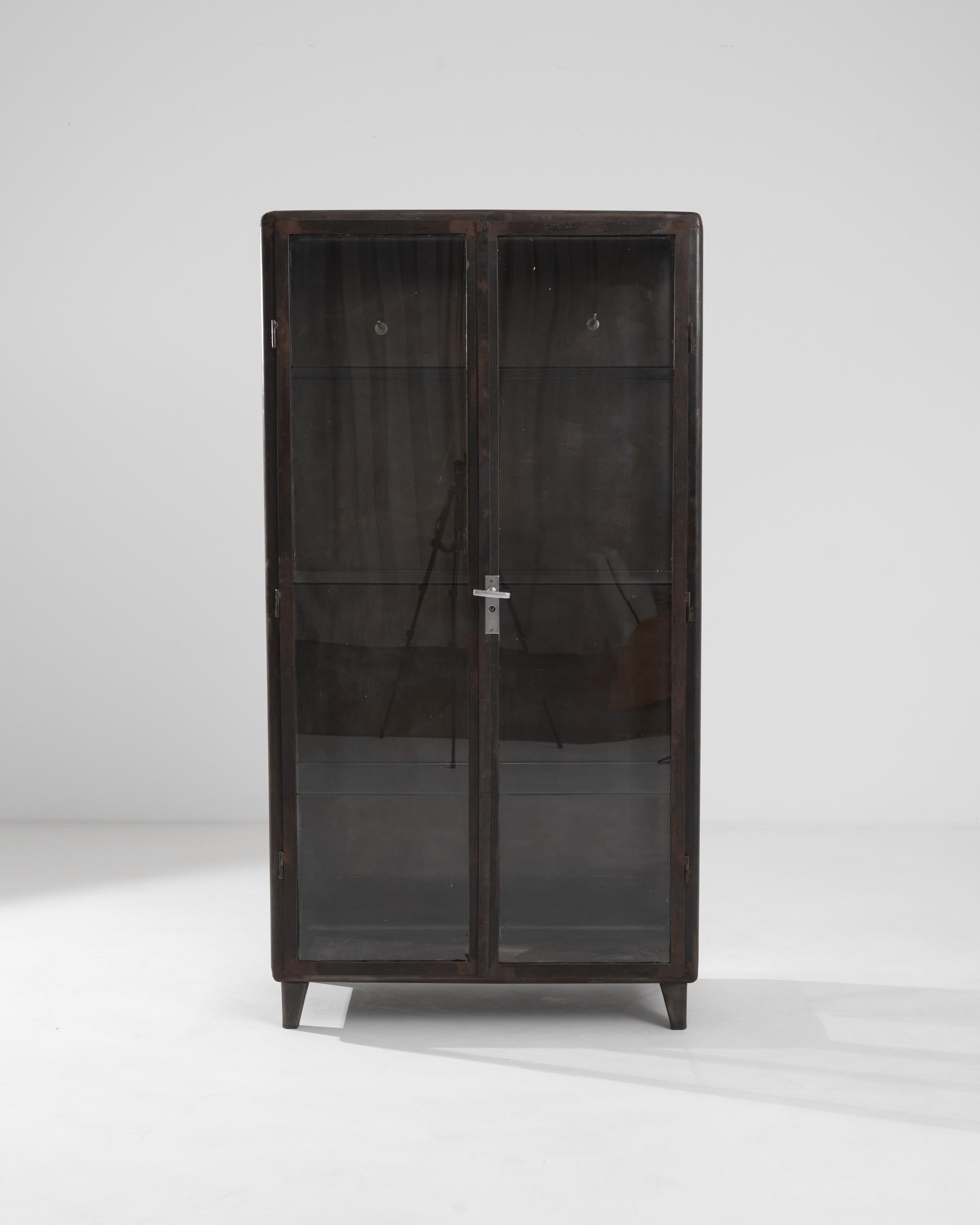 A metal display cabinet from 1950s Central Europe. Welded metal joinery, smoothed at the corners forms a utilitarian but eye-pleasing framing for this cabinet’s delicately composed glass shelves and drawers. Created with industrial mid-century
