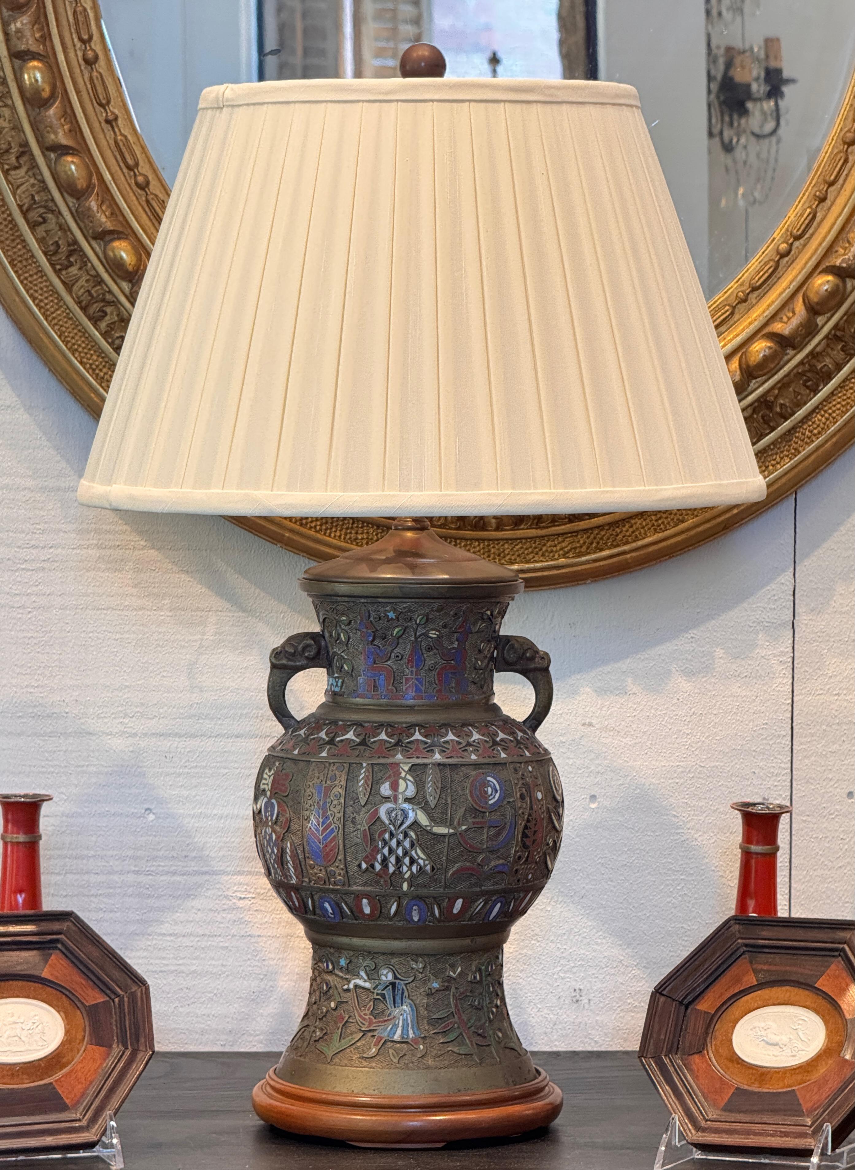 Lampe de table en bronze à décor émaillé champlevé coloré, présentant des motifs figuratifs et botaniques en rouge, blanc et bleu sur un fond finement texturé. Monté sur une base en bois tourné avec deux poignées à volutes à l'épaule.