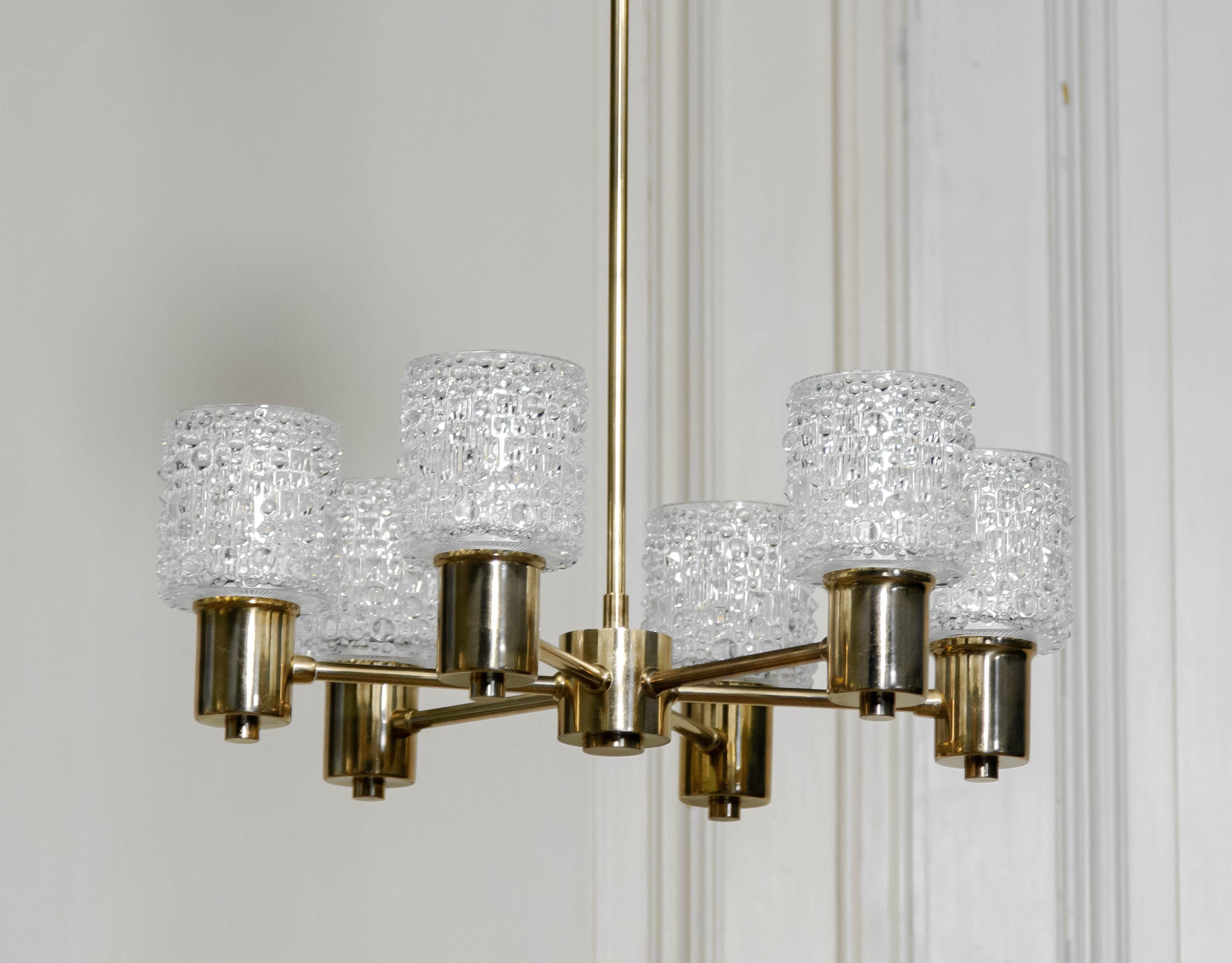 An elegant six-arm chandelier in solid brass, featuring textured Orrefors art glass shades. Each cylindrical glass shade is richly detailed with a bubbled surface, creating a sparkling effect when lit. The brass frame is geometrically arranged with