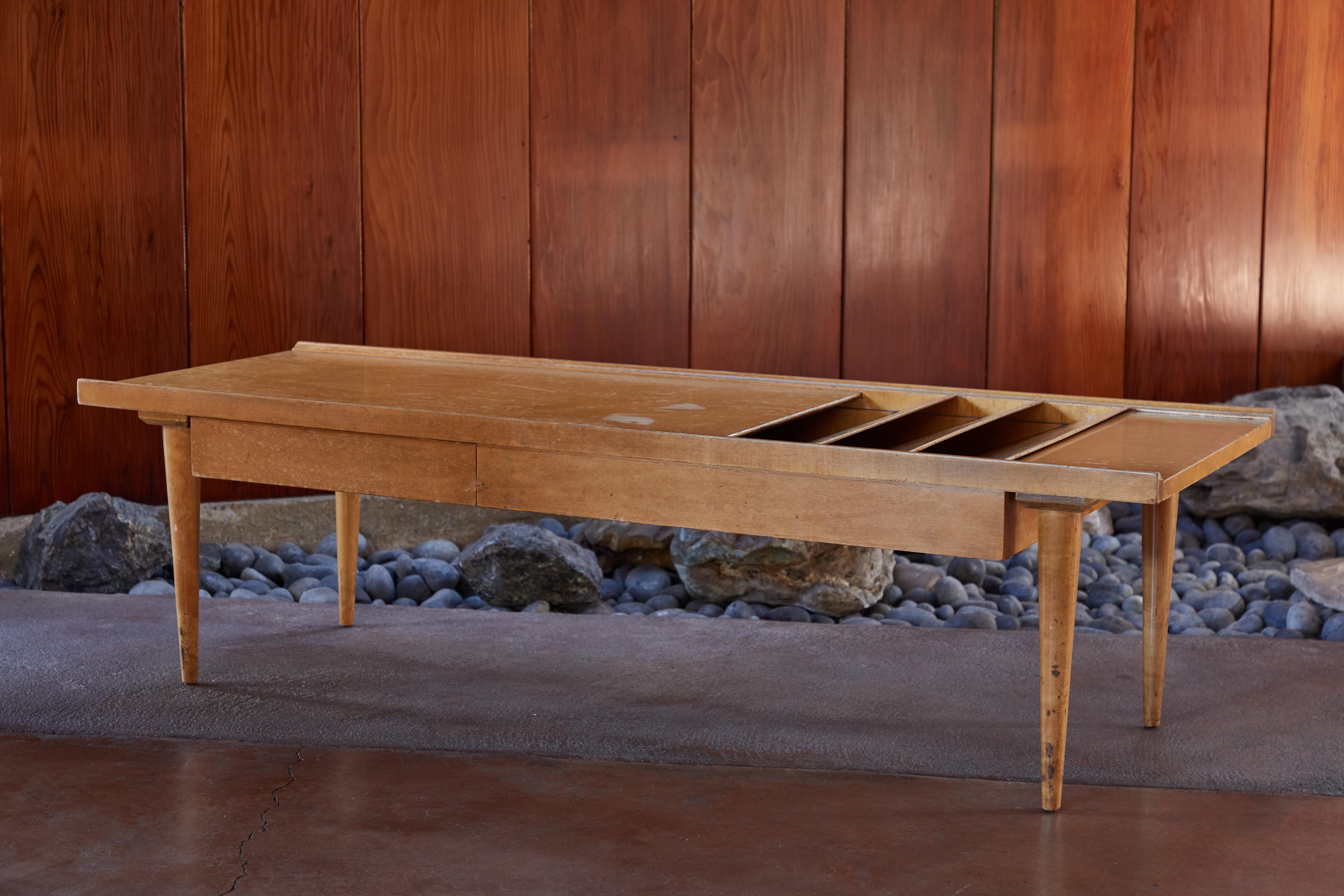 1950s coffee table in the style of Paul McCobb. A highly refined piece executed in blonde wood with magazine top file on one end and a side drawer on the opposite end. Supported by four signature angular tapered legs in a style suggestive of the