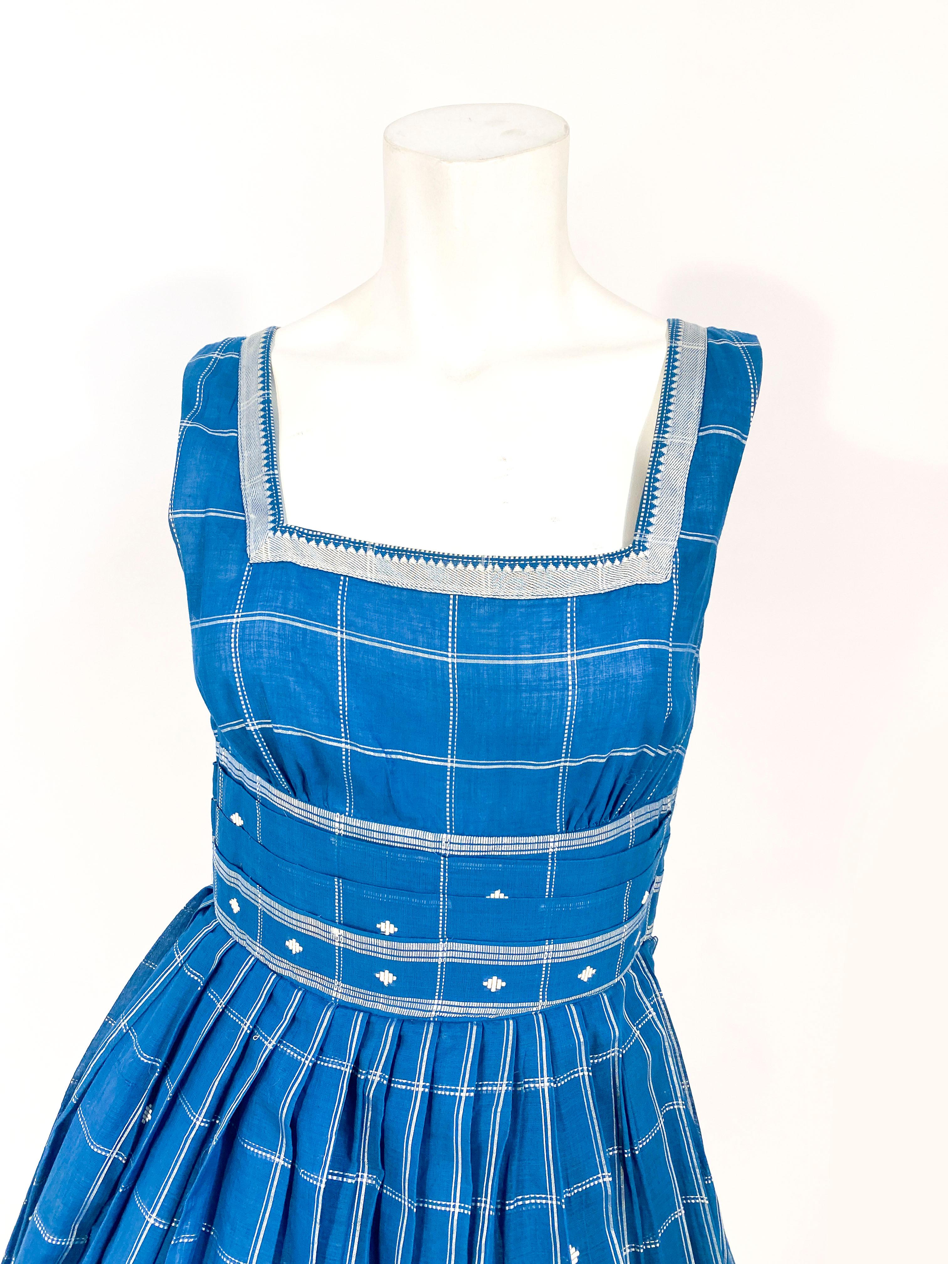 1950s custom-made cornflower blue day dress with a full pleated skirt, applied silver borders along the hem and square neckline, and a pleated waist. The textile has silver threading accents in plaid and small diamond shapes.